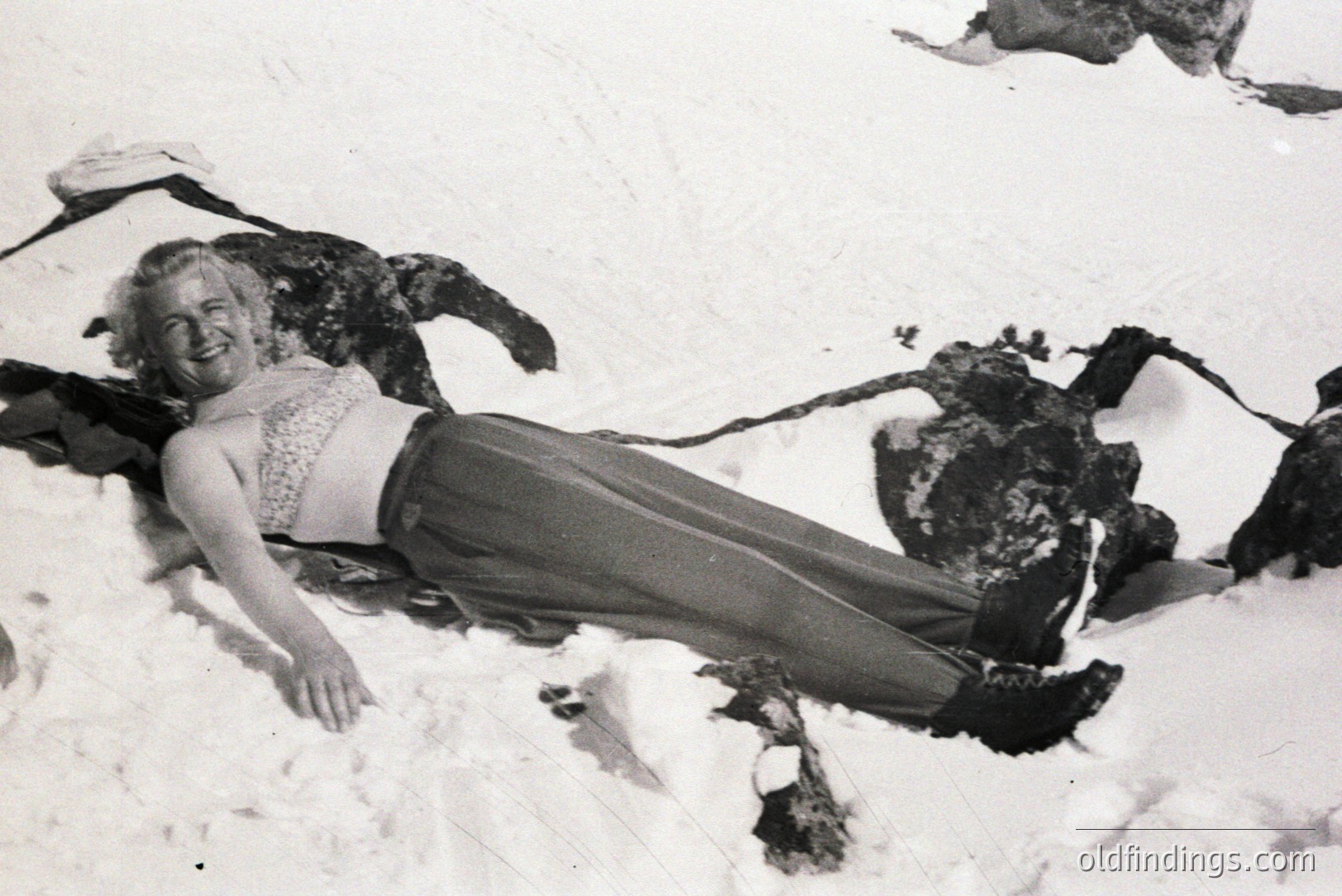 A smiling woman reclines in the snow, wearing a patterned bra top and wide-legged trousers. The image, likely taken in the 1960s or 70s, presents a playful, informal portrait. The snowy landscape, and the subject's relaxed pose, suggest a casual outdoor recreation. Could be used for vintage lifestyle or fashion stock.