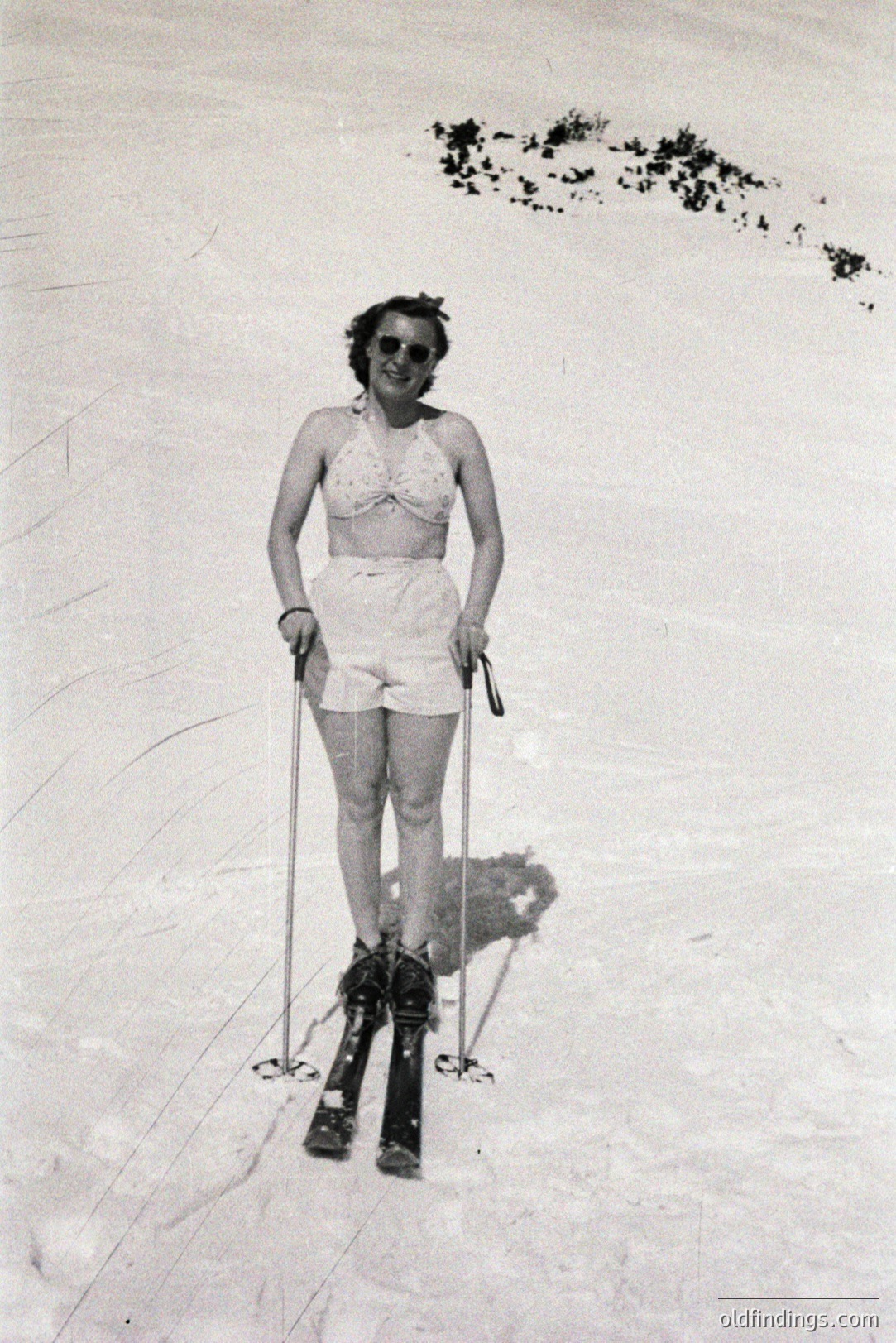 A woman in a vintage swimsuit and ski boots poses on snow. She wears sunglasses and holds ski poles, appearing poised. Likely a resort scene, potentially 1930s-1950s based on attire & photographic style. Historical record of winter recreation.