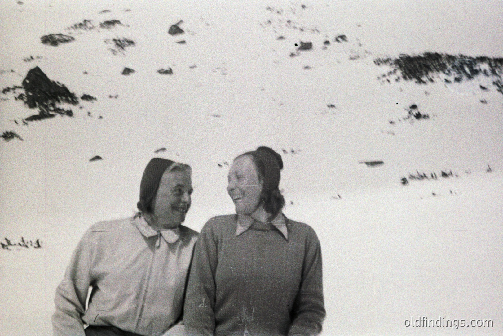 Two women with smiling expressions, bundled in warm winter clothing & hats, pose outdoors in a snowy landscape. Likely a candid moment, with a distant treeline visible. Appears to be a mid-20th Century (1940s-1960s) informal portrait.