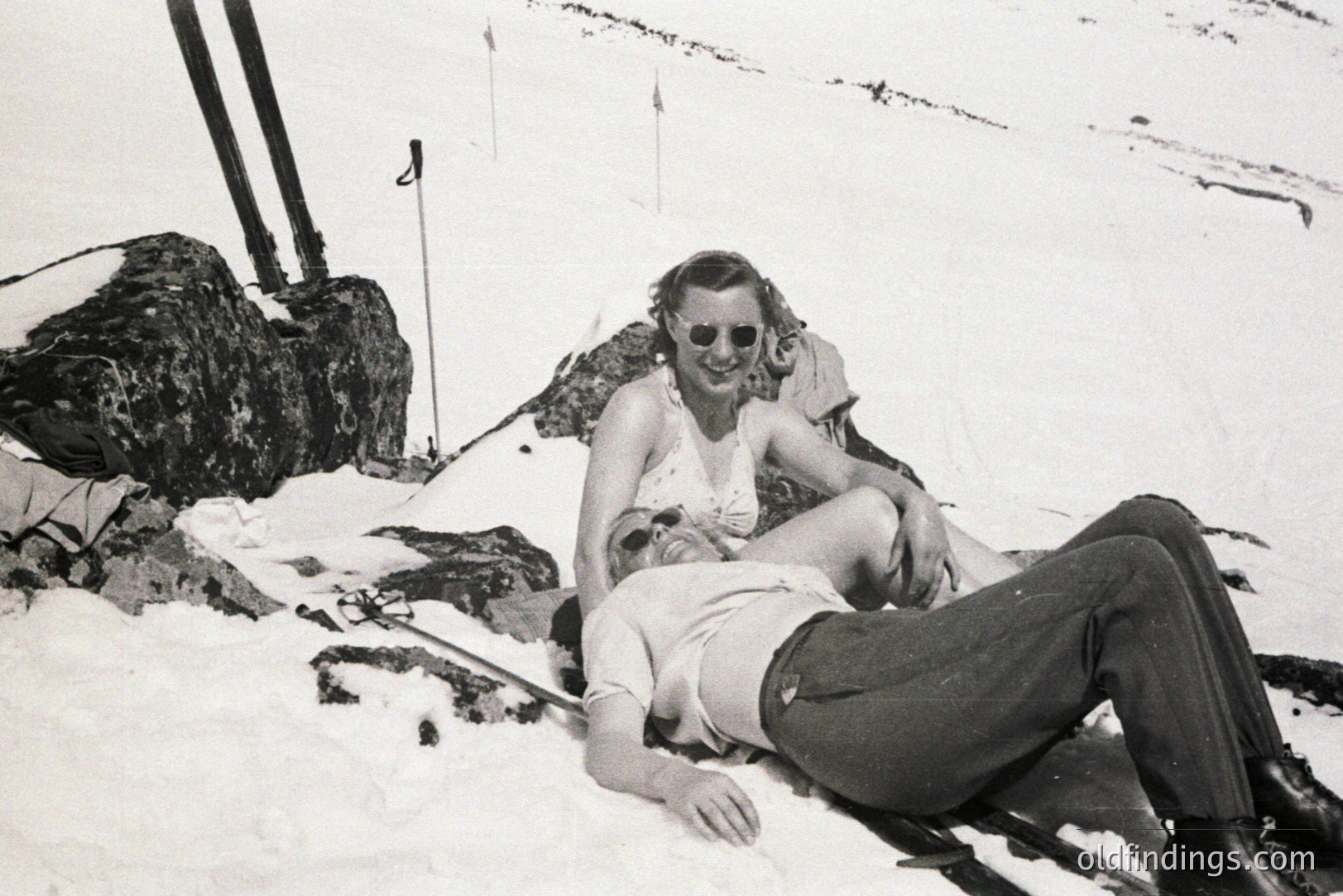 A man and woman rest on a snowy slope, reclined against a large rock formation. The woman, wearing a sleeveless dress and sunglasses, sits atop the man who is lying flat. Ski poles and a snow fence are visible in the background. Likely a leisure activity scene, circa 1930s-1950s.