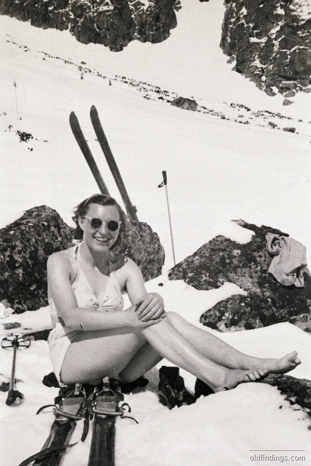 A poised woman in a one-piece swimsuit sits on a snowbank, legs crossed, with vintage skis and poles nearby. She wears round sunglasses. Likely 1960s alpine leisure; a stylish, confident representation of outdoor recreation. Location uncertain, but appears to be a mountainous region. The image exhibits strong commercial appeal for vintage lifestyle design.