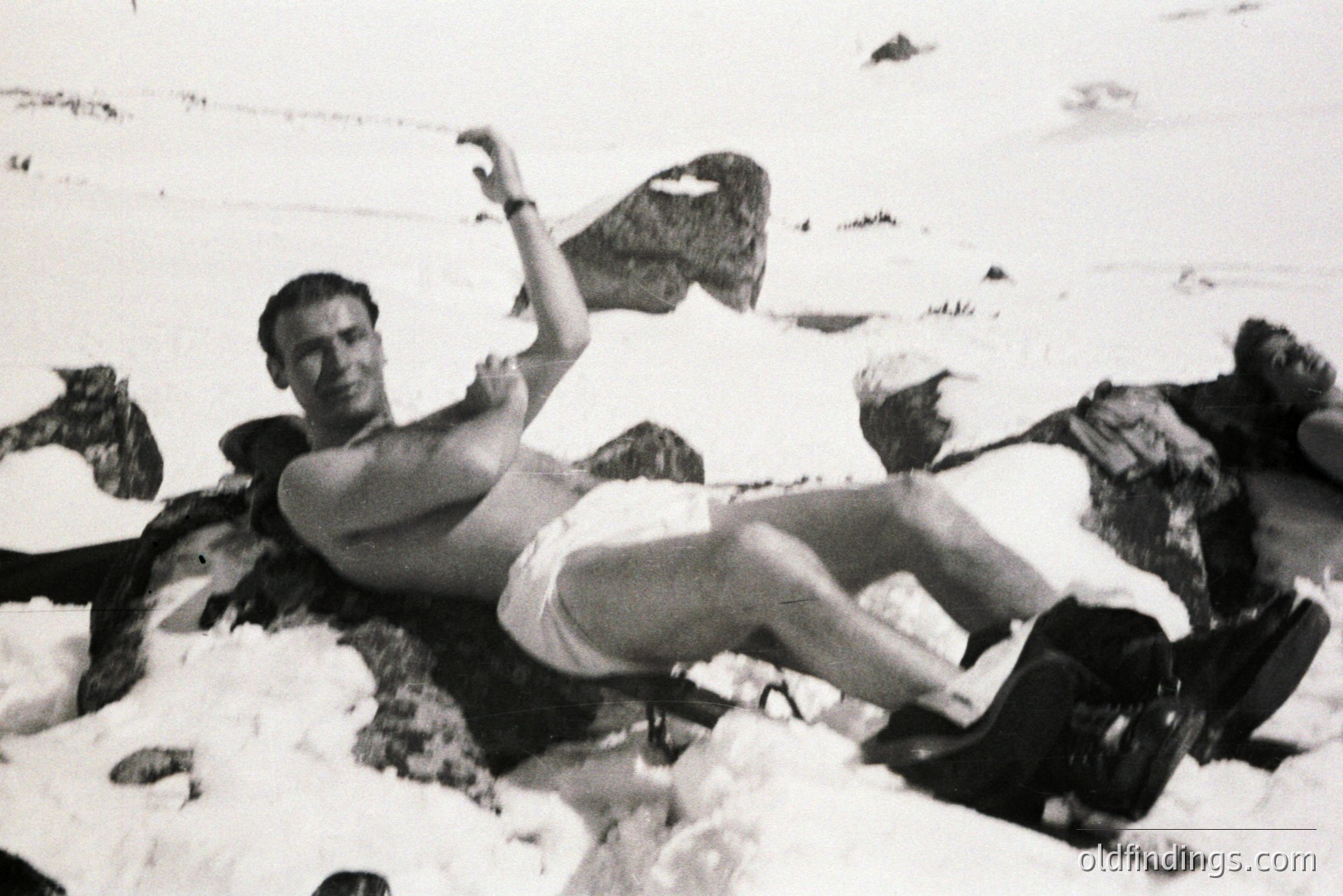 A man, shirtless and in shorts, reclines on a snow-covered rock formation. He appears to be posing with an air of relaxed confidence. A woman stands in the background, partially obscured. Likely a promotional or lifestyle image, possibly for outdoor apparel.