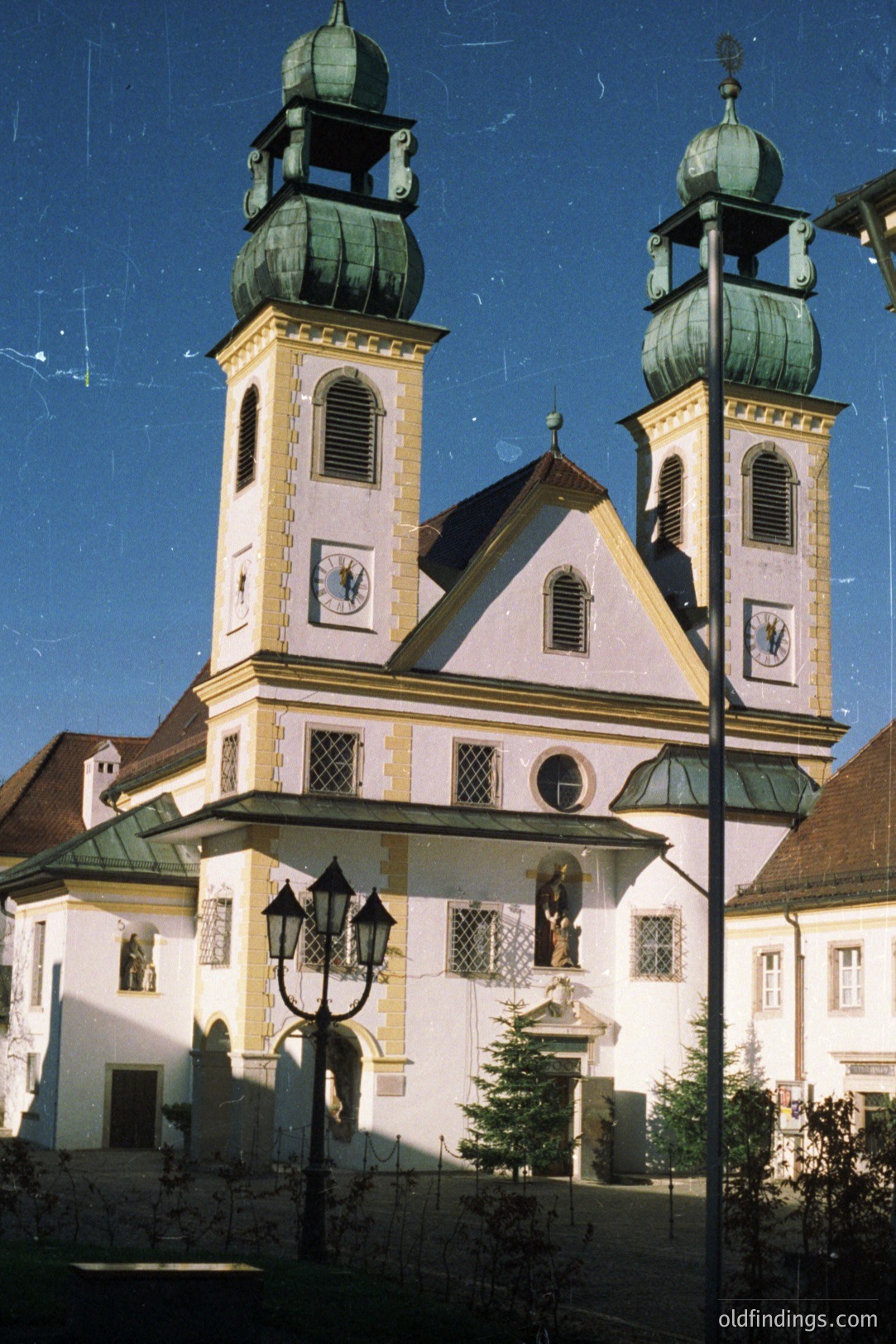 Grand Baroque-style church facade features twin towers topped with green domes. The structure includes intricate detailing, symmetrical design, and a prominent central entrance with a statue. Likely central European, possibly 18th century. Excellent architectural reference.