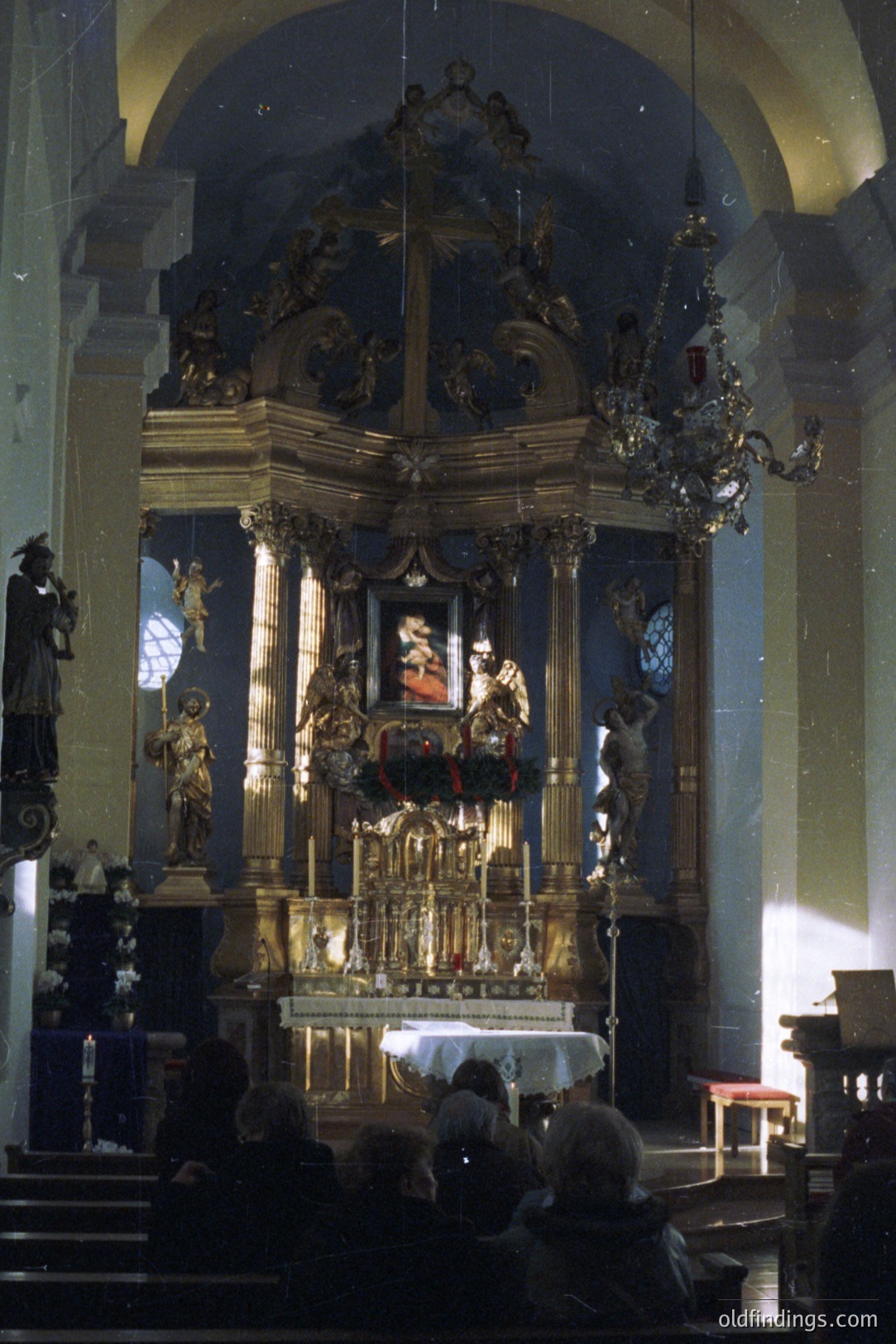 Ornate gilded altar dominates a church interior. A depiction of a figure is centered within, flanked by statues and elaborate detailing. Visible attendees are blurred in the foreground. Likely mid-20th century, potentially European.