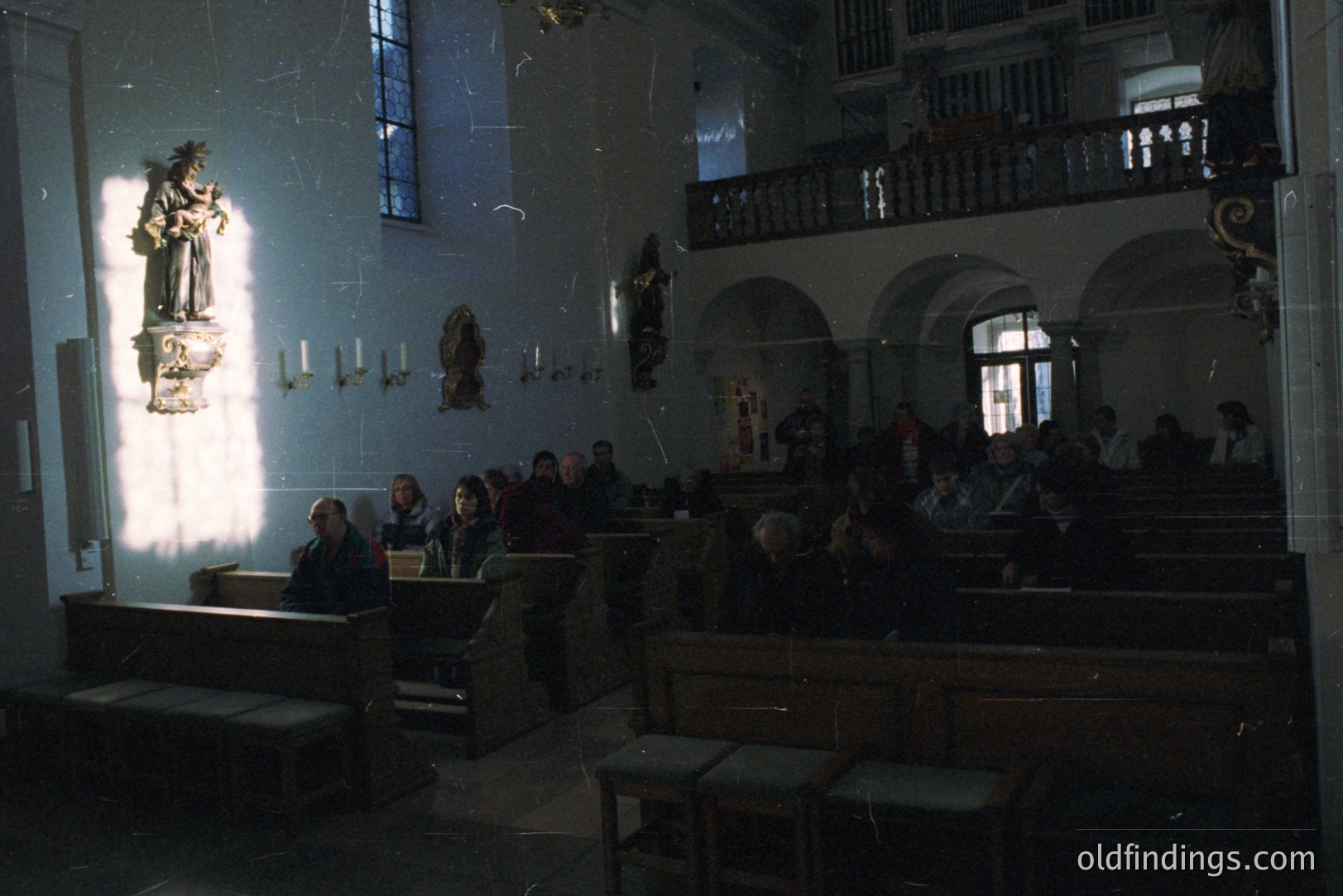 A view inside a church. Several people are seated in pews, observing an event or service. Stone statues and architectural details adorn the walls and upper levels. Visible sunlight streams through a window, illuminating the scene. Likely mid-20th century.