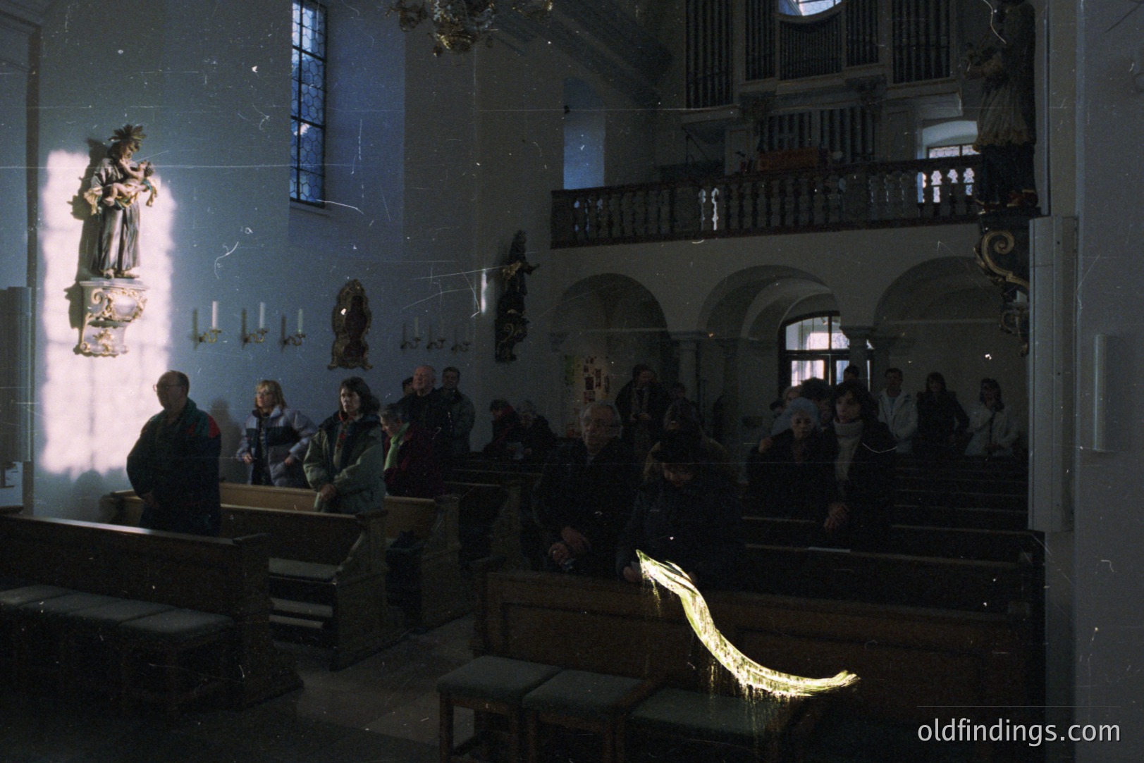 A dimly lit, historic church interior. Several figures are seated in pews while others stand or are visible on a balcony. A statue is illuminated by a strong light source from a window. Architectural details include arched doorways, a pipe organ, and decorative elements on the balcony. Likely 1970s or 80s, based on clothing styles.