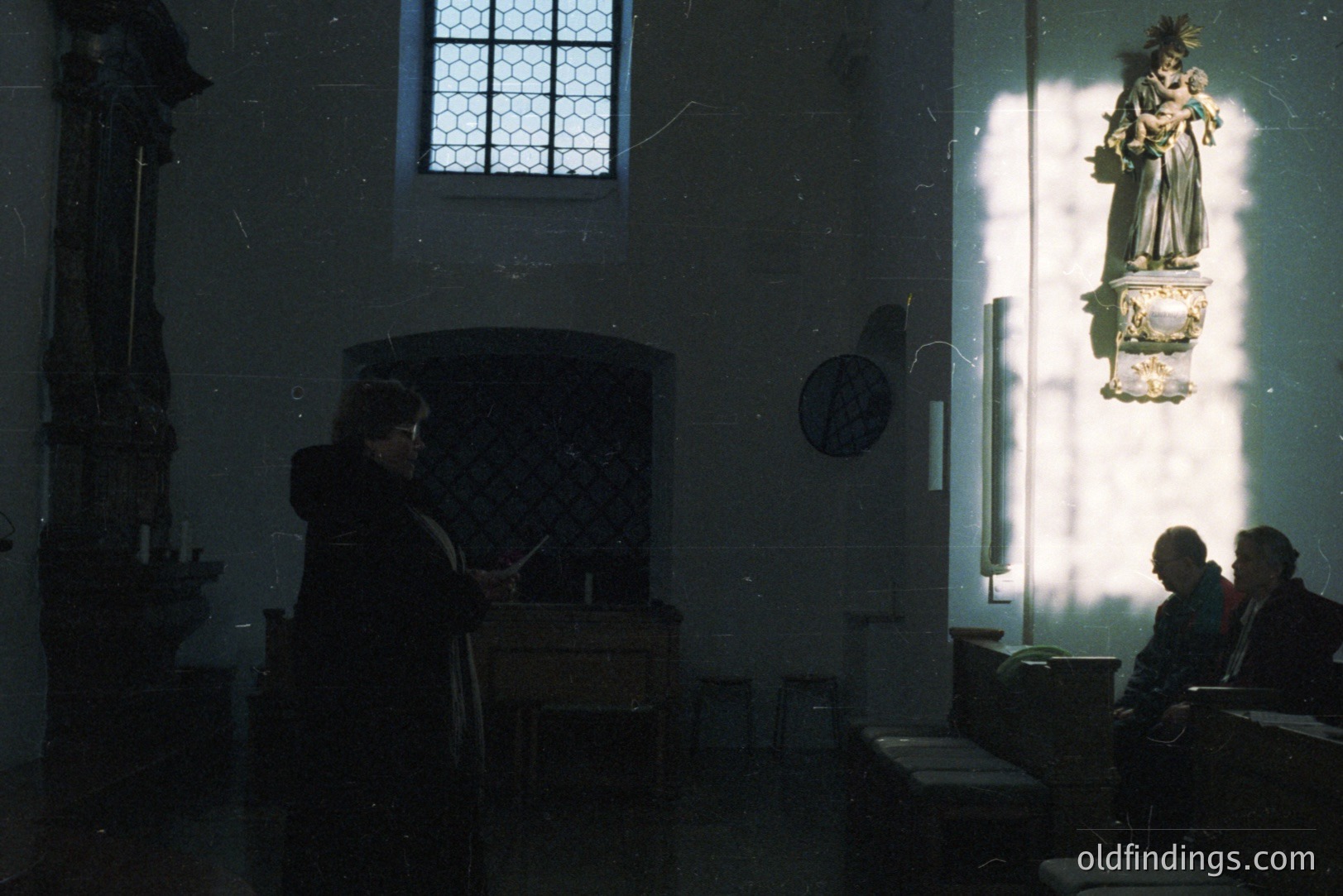 Interior of a stone church, featuring a dark wooden pulpit, simple benches, and a statue of a robed figure illuminated by sunlight streaming through a stained-glass window. Two figures are visible - one near the pulpit, one seated. Appears to be a moment of quiet contemplation.