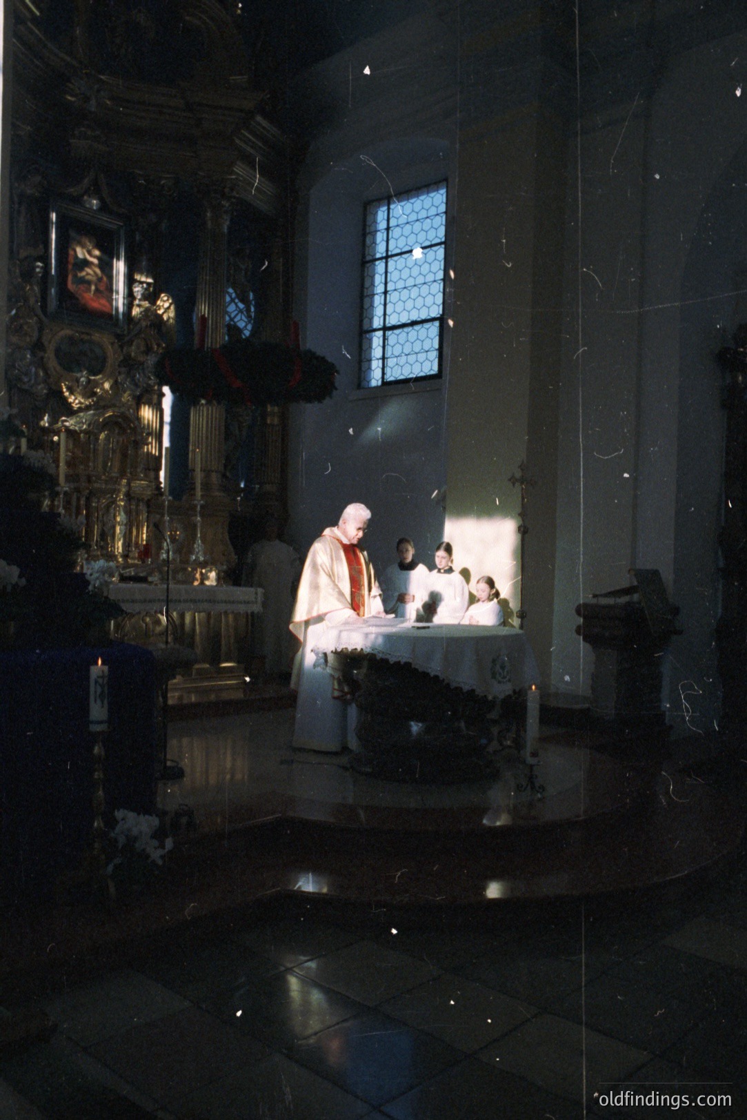 A solemn religious scene within a Catholic church. A priest in vestments presides over the altar, with acolytes nearby. Ornate, gilded baroque detailing adorns the altar backdrop and a side window casts a dramatic light shaft. Appears to be from the late 20th century.