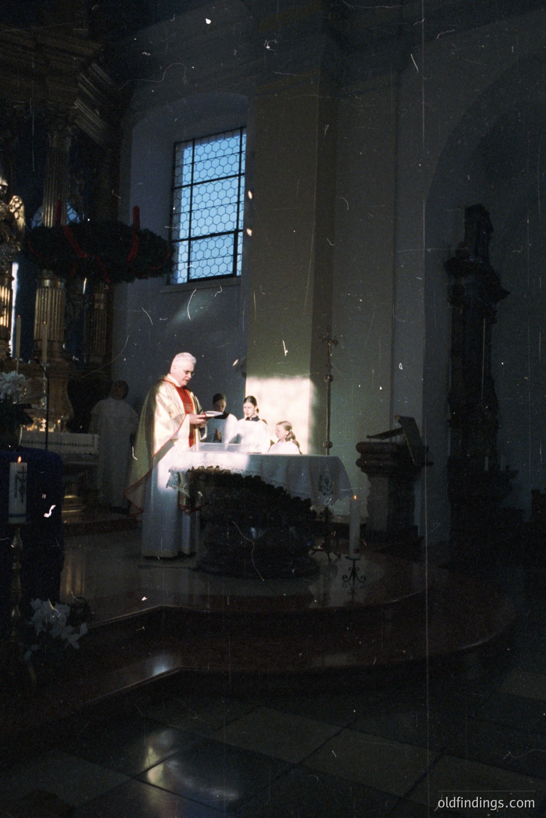 A Roman Catholic Mass is underway, featuring a priest administering communion. The interior showcases ornate architecture, including a stained-glass window and stone carvings. Lighting suggests a 1960s-1970s era, possibly European. Likely a stock photography reference for religious themes.