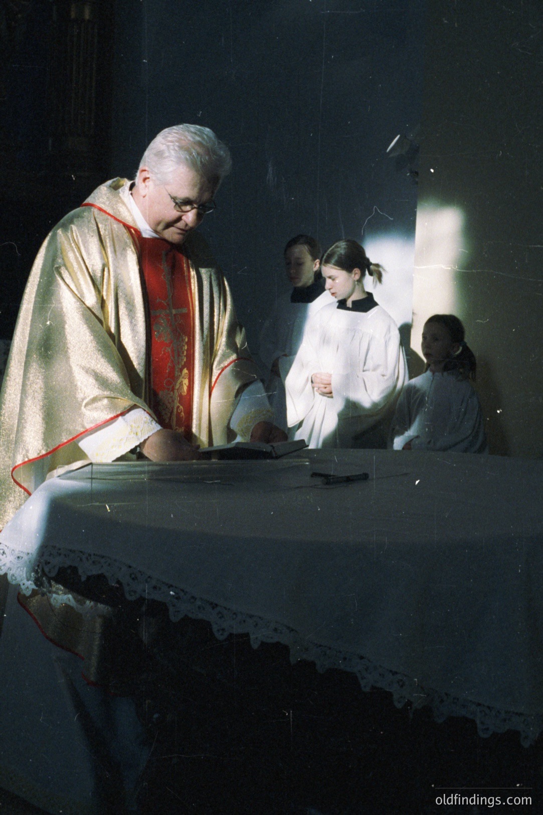 A Roman Catholic priest in ornate vestments oversees a service, seemingly reading from a book on an altar draped in white lace. Two young altar servers, also in white robes, stand attentively. Soft, diffused light highlights the scene, creating a sense of reverence. Likely 1970s-80s.