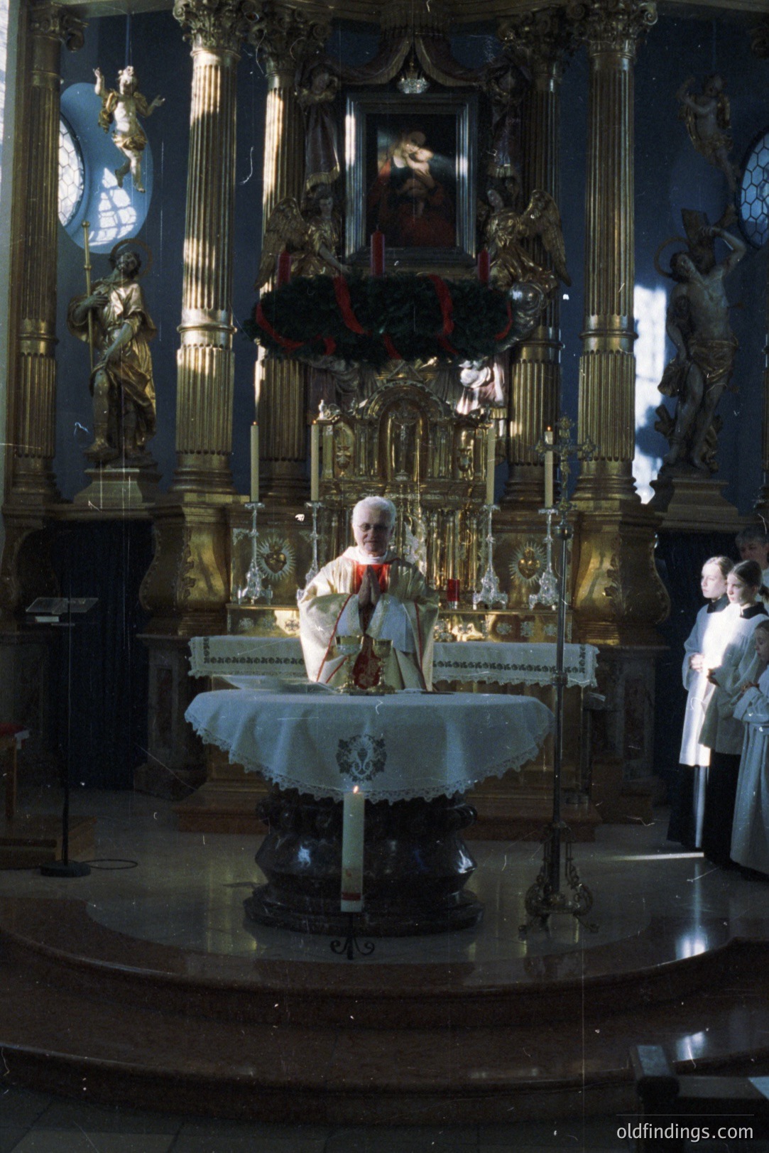 A Catholic priest, likely celebrating mass, stands behind an ornate altar with gilded carvings. Statues of angels flank the scene, bathed in light from unseen windows. The altar is draped with a white cloth, adorned with candles and a floral arrangement. Several robed figures stand observing. Likely mid-20th century.