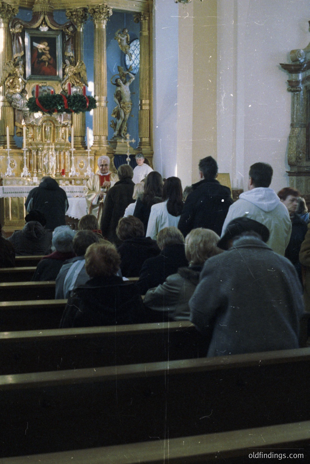 A formal religious service unfolds within a traditional Catholic church. Attendees are seated in pews, facing an ornate altar and intricately carved reredos featuring a crucifix. The scene suggests a confirmation or similar rite of passage, given the presence of individuals in white robes. Likely 1970s-1980s, color film.
