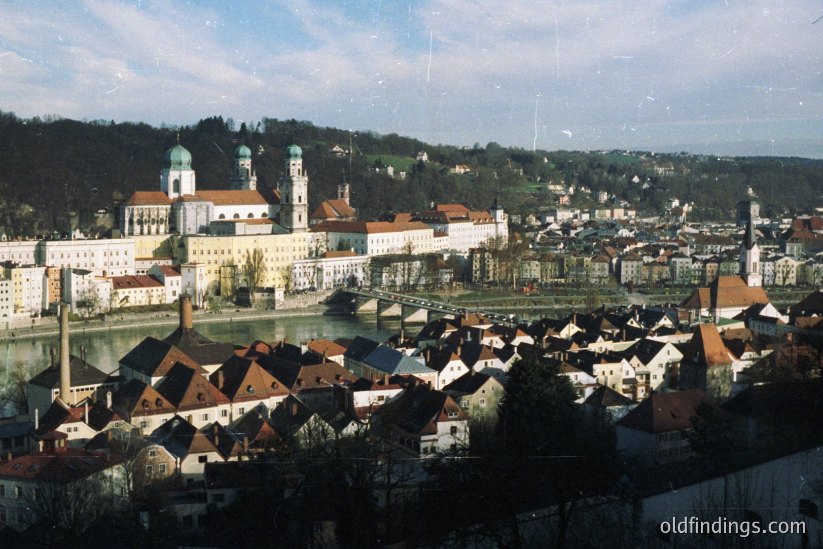 A panoramic view of the historic town of Passau, Germany, showcasing a riverfront cityscape with red-tiled roofs and the impressive St. Stephan’s Cathedral dominating the skyline. Likely taken during the late 20th century, architectural details suggest traditional Bavarian style. A bridge spans the river, connecting the town’s sections.