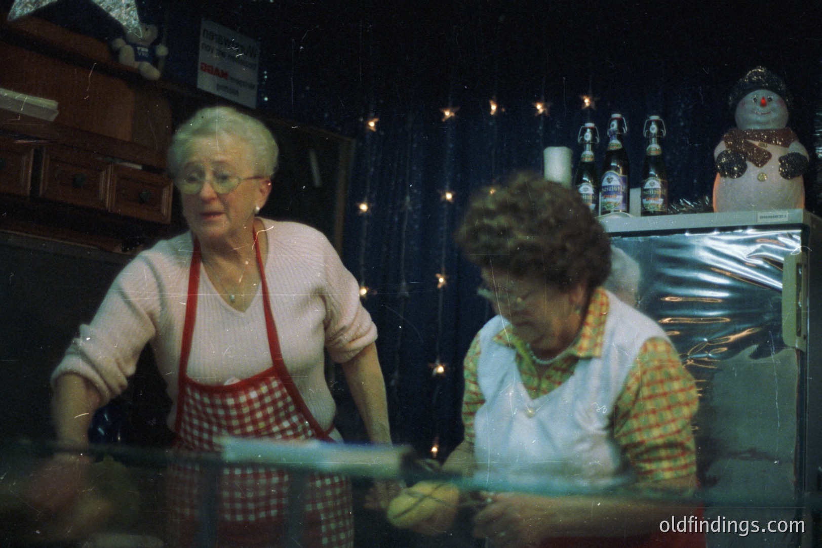 Two women in aprons, likely shopkeepers, work behind a display counter filled with candies. Festive decorations, including a snowman and string lights, adorn the space. Likely a holiday-themed confectionery shop scene, aesthetic. Professional photography or stock imagery potential.