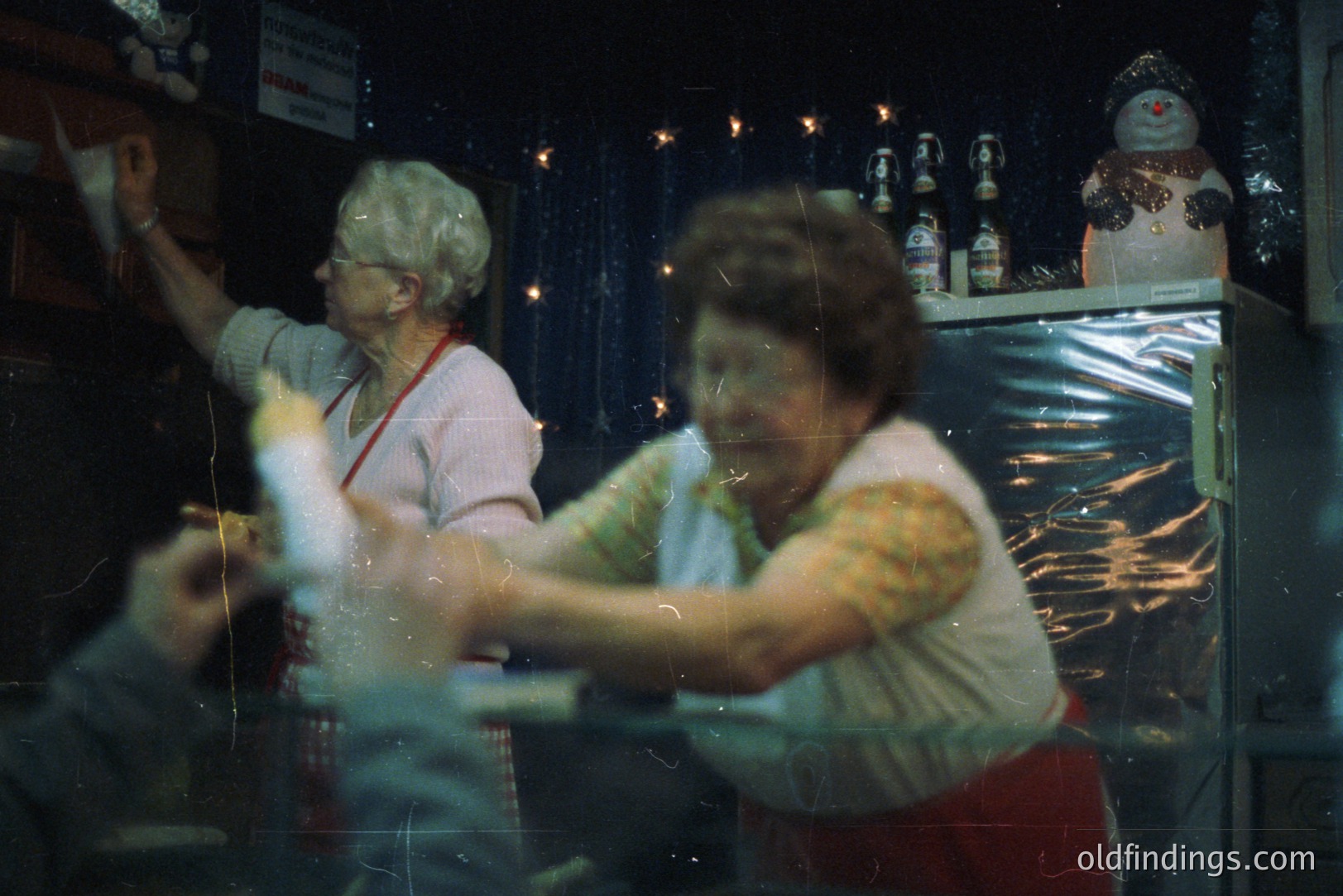 Two women in aprons work behind a counter, likely in a confectionery or gift shop. Visible merchandise includes figurines, decorations, and glassware. The image has a slightly blurred, vintage aesthetic. Likely taken in the 1970s or 1980s.