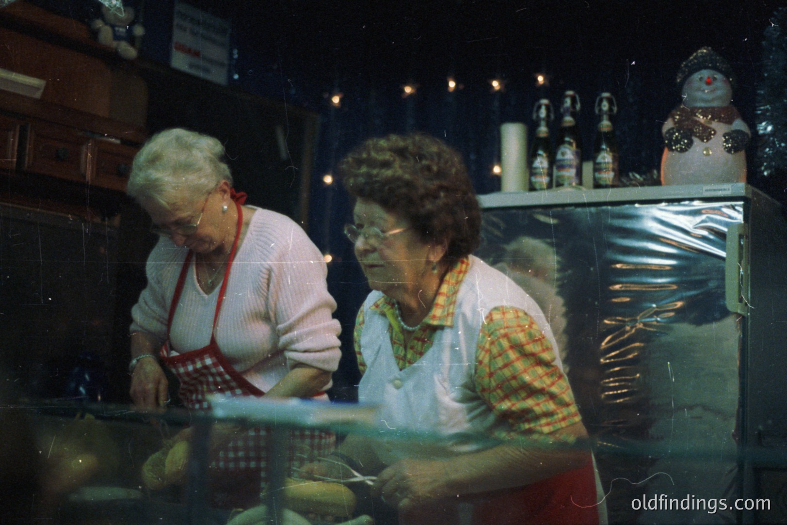 Two women in work aprons prepare food behind a stainless steel counter. A display of ornaments and decorations hangs on the wall. Likely a festive market or shop scene. Appears to be a snapshot from the 1970s or 80s. Potential stock photo reference for vintage Christmas/food prep.