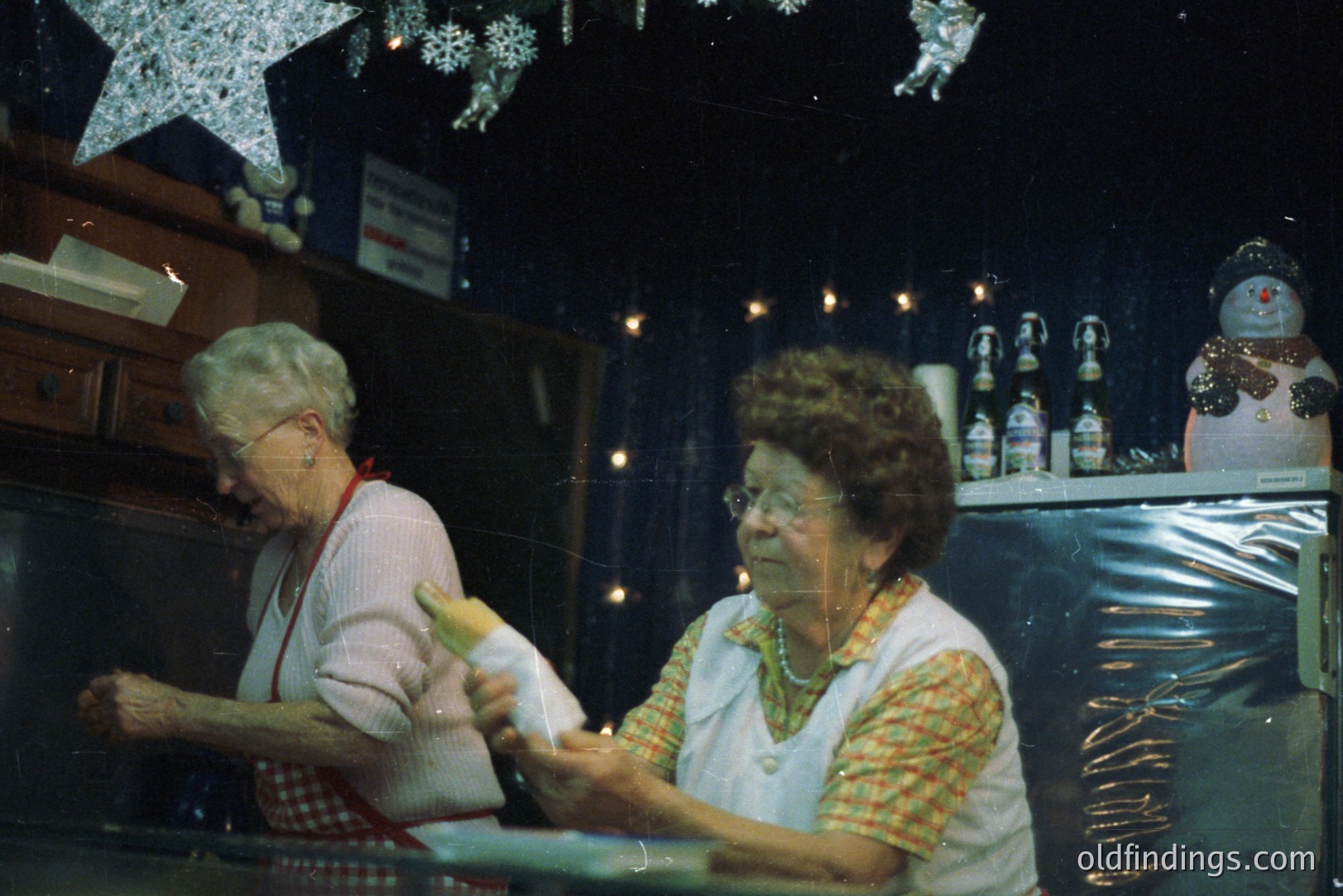 Two older women in aprons work behind a counter, likely preparing food or beverages. Christmas decorations – a snowman, twinkling lights, and a starburst – adorn the background. Likely a small business or community event, possibly the 1970s.