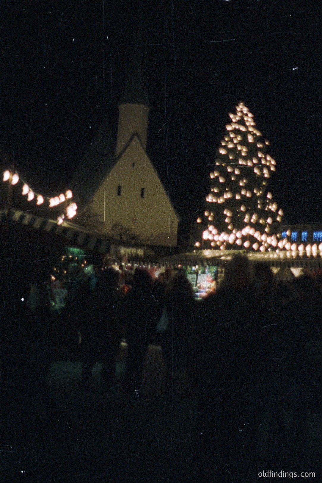 A community gathers at a nighttime Christmas market. A simple, peaked-roof building flanks a brightly lit, artificial fir tree. Festive string lights adorn the market stalls. Possible 1970s/80s snapshot captures a seasonal gathering. Ideal for design referencing and nostalgic stock imagery.