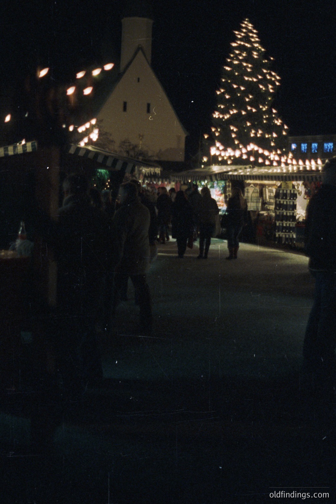 A brightly lit, snow-covered outdoor ice rink with a large, decorated Christmas tree. Figures in winter clothing skate around, viewed from a slightly elevated perspective. Appears to be a festive, likely European, winter scene. Likely 1970s-1980s film photography. Could be a commercial stock image depicting holiday atmosphere.