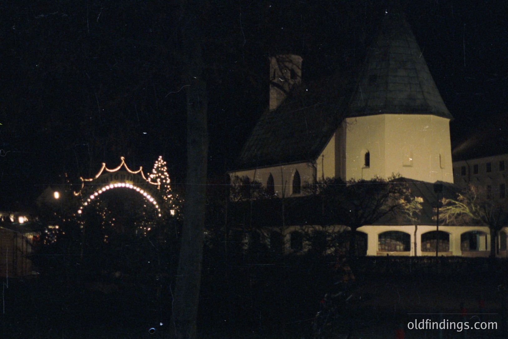 Night view of a stone building with a distinctive, domed tower and arched entrance decorated with string lights. Likely a European castle or historic structure. Appears to be a festive event or holiday display. Location and specific date unknown.