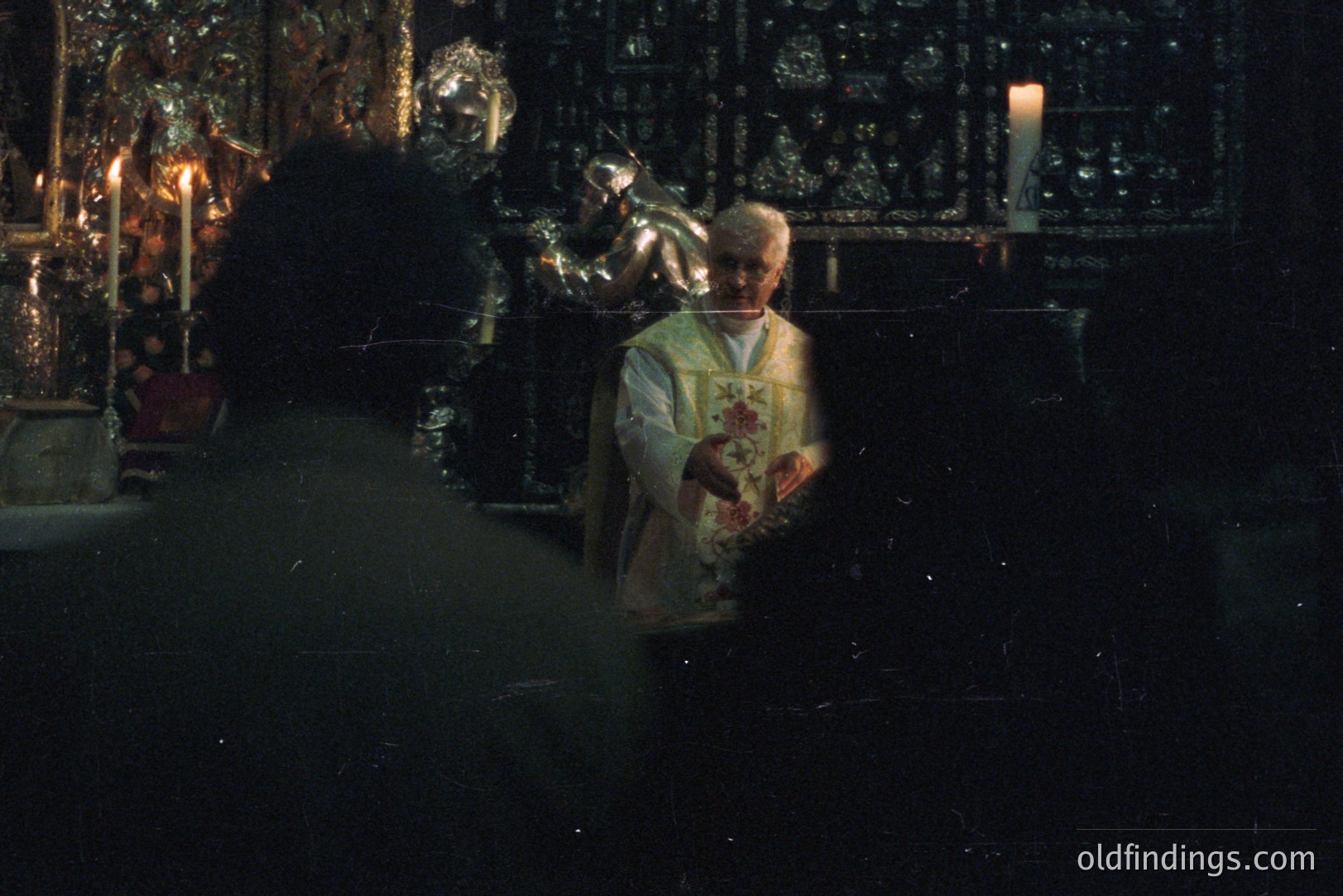 A dimly lit scene depicts a man in a white ecclesiastical robe administering communion. He holds a chalice, facing a partially visible figure in shadow. Candles illuminate the foreground and ornate, reflective altar pieces. Likely a religious ceremony, possibly Catholic.