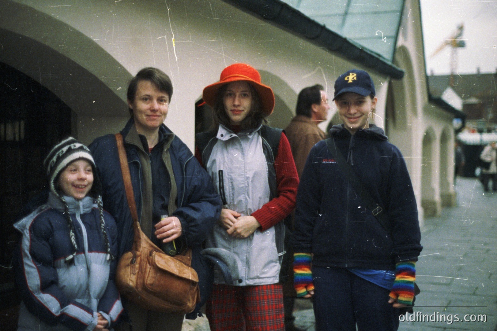 A family of four stands in front of a historic, arched building. The subjects are wearing 1980s/90s outerwear and accessories. The building architecture suggests a European locale. A vintage camera bag hangs from the woman’s shoulder.