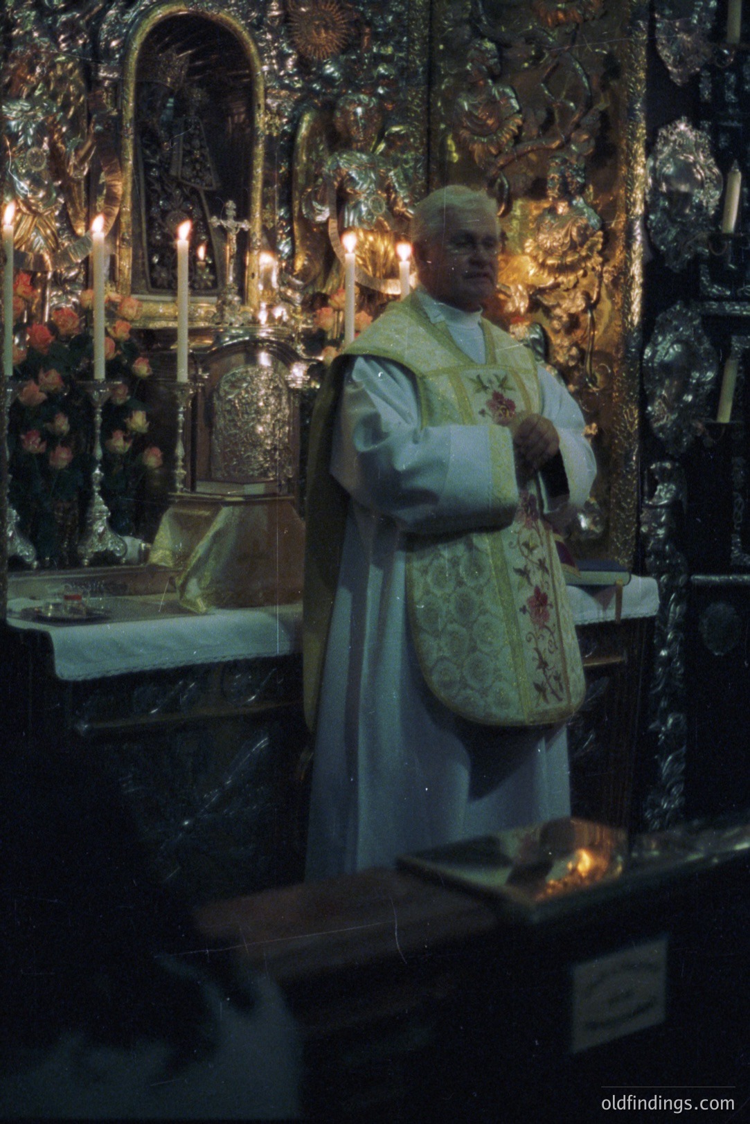A Roman Catholic priest, wearing ornate vestments, stands before an elaborately carved and gilded altar, illuminated by numerous candles. The scene conveys a sense of traditional religious ceremony. Likely from the mid-20th century, possibly Eastern Europe or Italy. A valuable historical document of faith.
