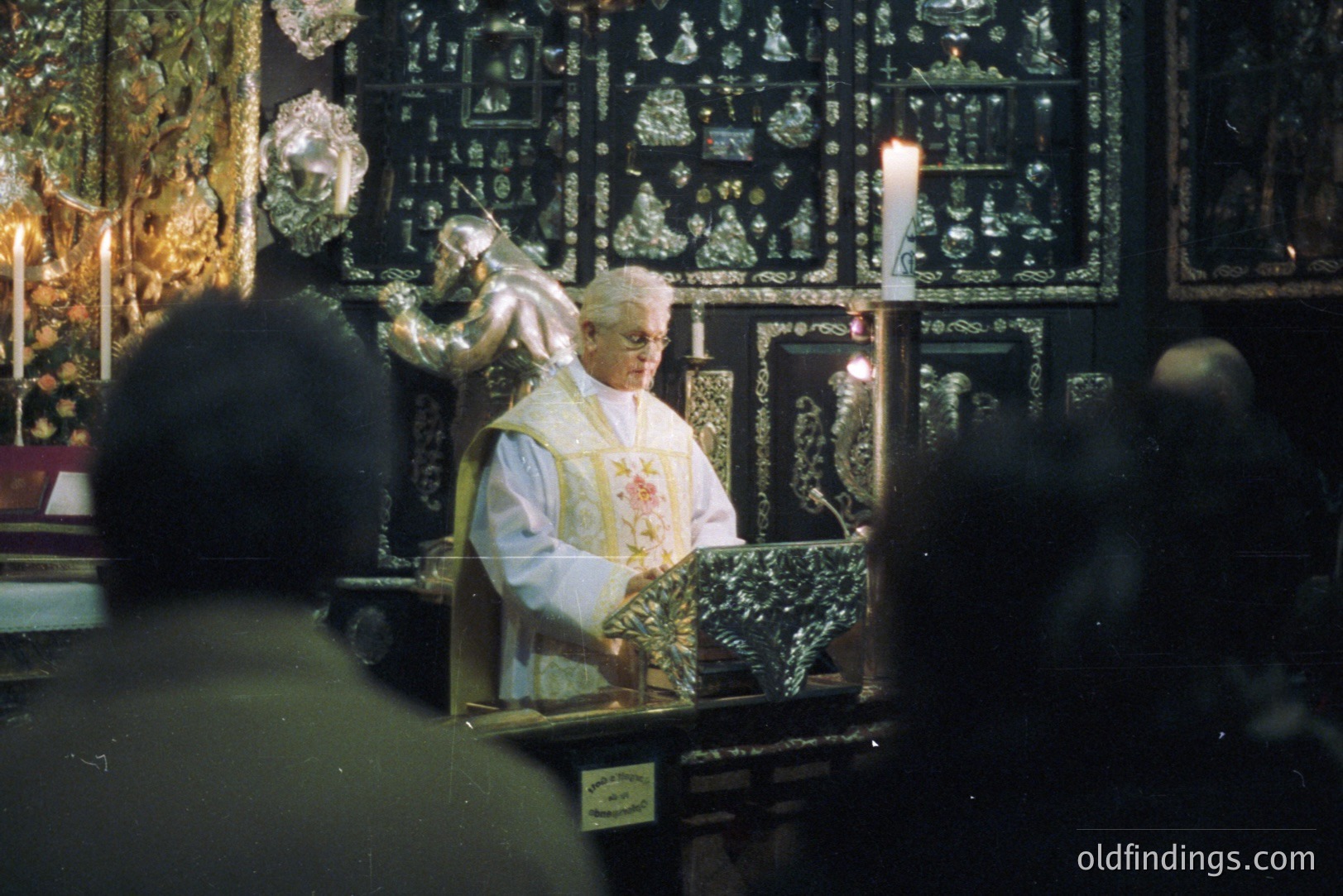A priest in vestments performs a ceremony behind an ornate, gilded altar screen. Details suggest a Catholic service. The style of the screen and the priest's appearance indicate a possible 1970s timeframe. Low-light conditions, typical of religious spaces. The image is cropped, showing the backs of attendees.
