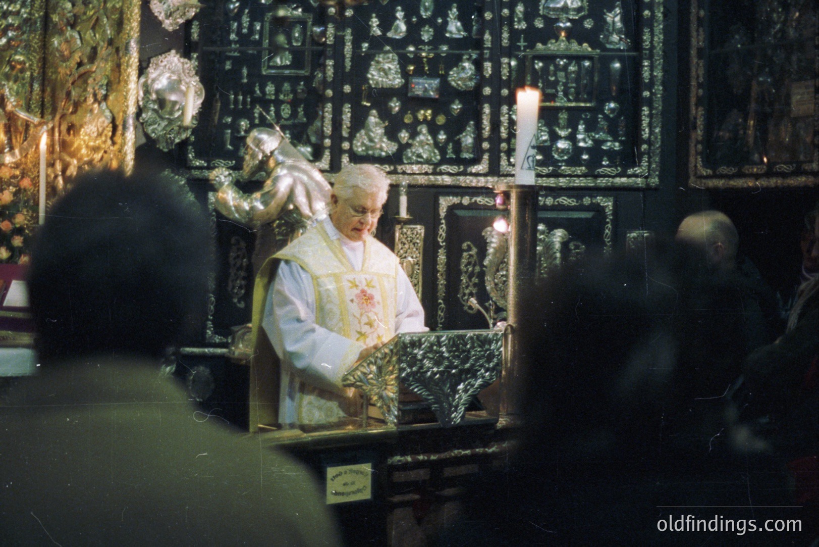 A priest in white robes presides over an ornate, gilded altar, likely during a religious ceremony. Candlelight illuminates the scene, enhancing the rich details of the altar's intricate carvings. Several figures are partially visible in the foreground. Likely 1970s or 80s film photograph.
