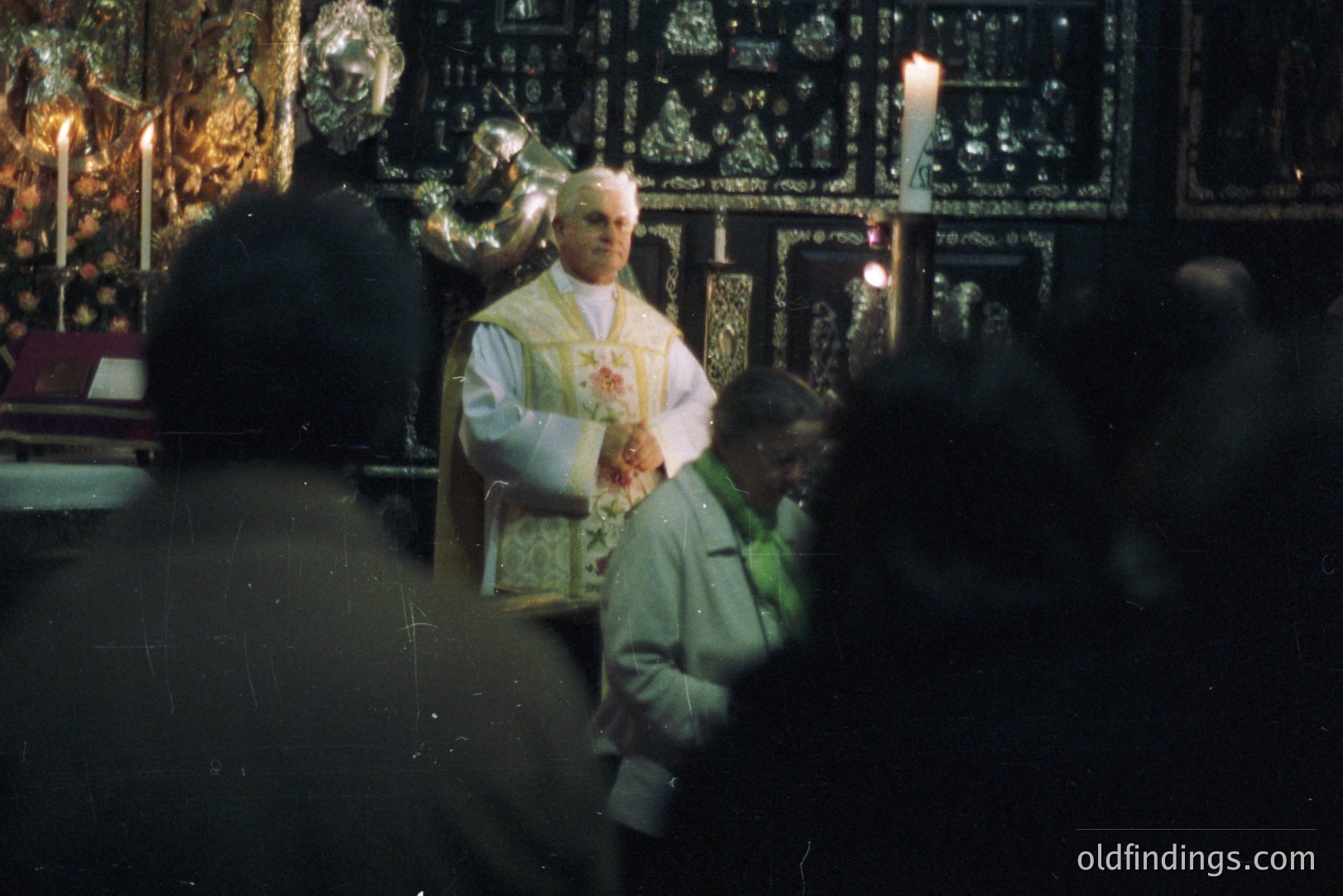 A Catholic priest in vestments elevates a host during a ceremony. Intricate, gilded altar decorations and flickering candles fill the background, visible through a crowd of attendees. Likely a liturgical photograph from the 1960s or 1970s, potentially a travel or religious stock image.