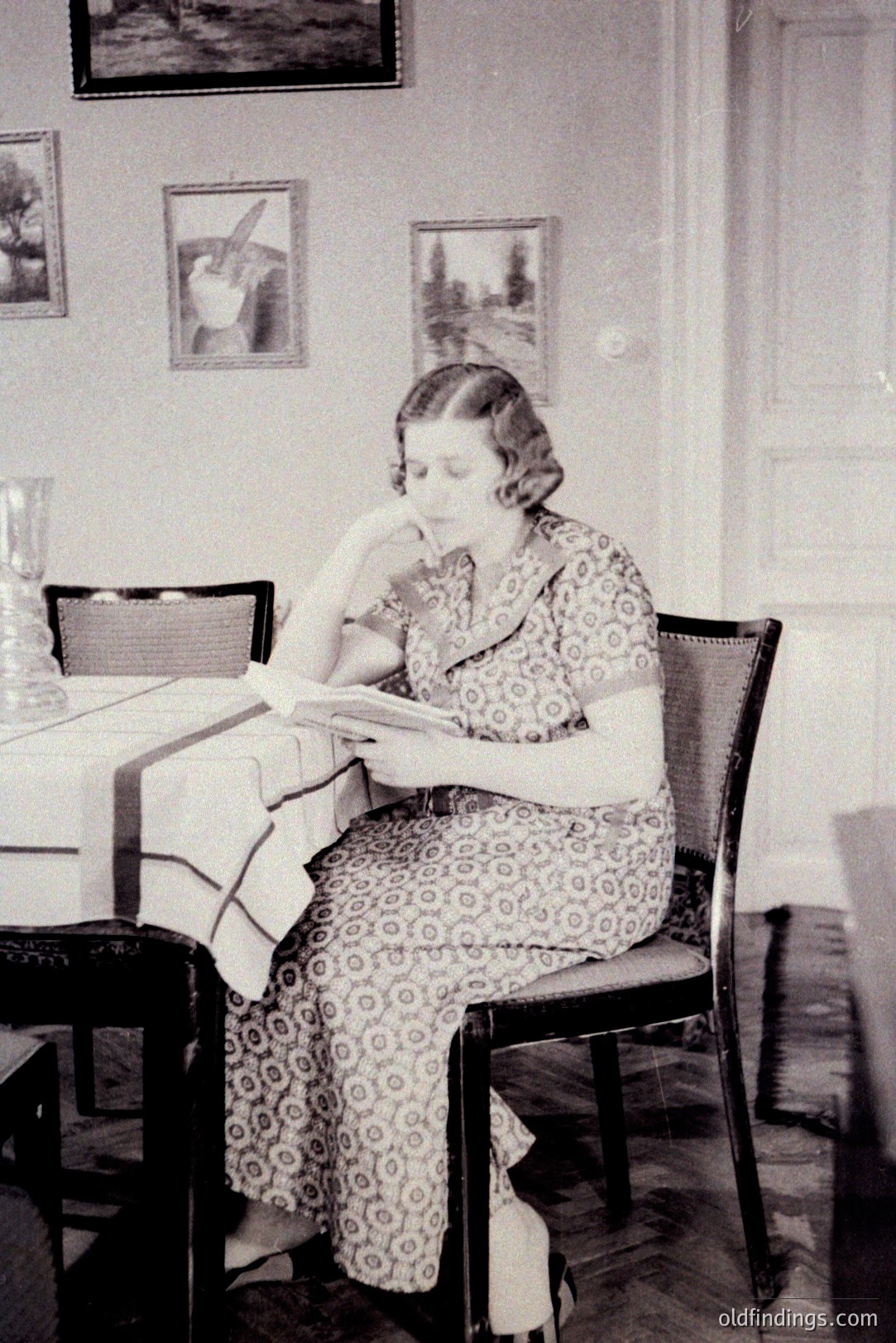 A woman seated at a table, engrossed in reading. She wears a patterned, long-sleeved dress and appears contemplative. A patterned tablecloth covers the table; framed artwork decorates the background wall. Likely a domestic interior, c. 1930s-1950s.