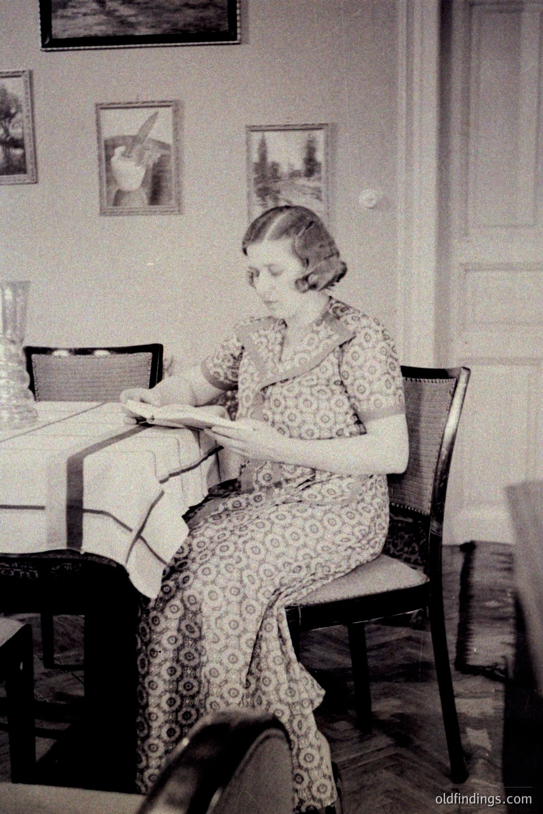 A woman sits at a table covered with fabric, intently examining a piece of paper or fabric. Interior scene with framed artwork on the walls and detailed parquet flooring. Likely 1920s-1930s, possibly European. A glimpse into domestic life.
