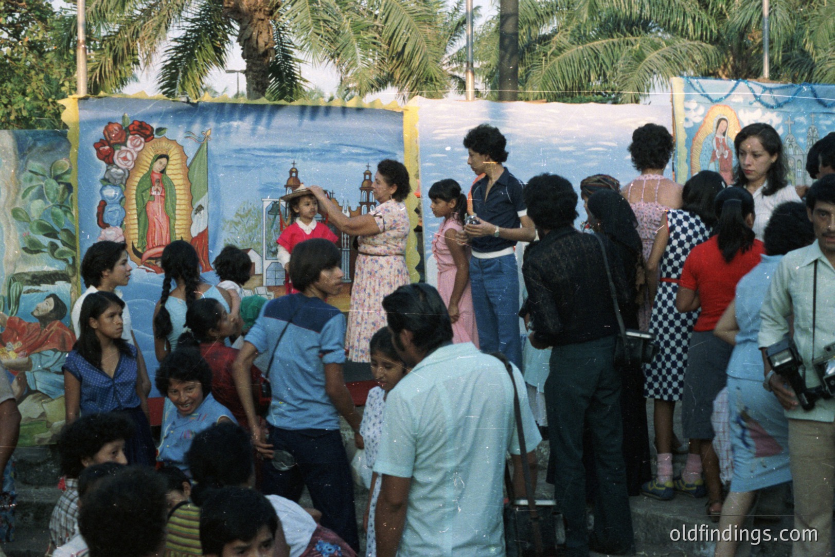 A vibrant scene: a crowd gathers before murals of the Virgin Mary. A woman in a floral dress raises a religious icon, while onlookers watch. Visible 1970s attire & hairstyles suggest a community gathering. Palm trees frame the background.