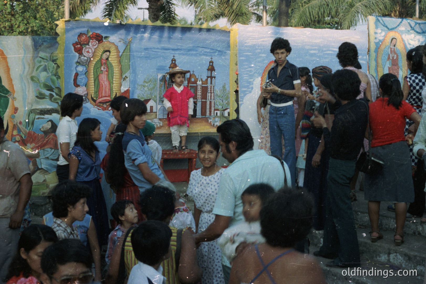 A young boy in a pink outfit stands on a small platform before a painted mural depicting religious figures and a town square. A crowd of people, typical of the 1970s, watches. Bell-bottoms, floral prints, and bowl haircuts are visible. Likely a local festival or celebration.