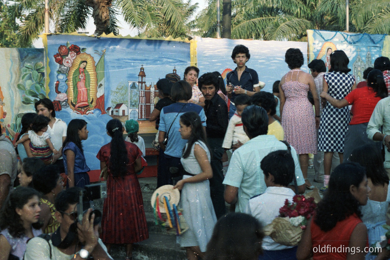 A vibrant outdoor gathering features a large mural of the Virgin of Guadalupe. People in 1970s attire—floral dresses, bell-bottoms—congregate; some hold flowers. A formal event, likely a religious festival, shown in a tropical location.