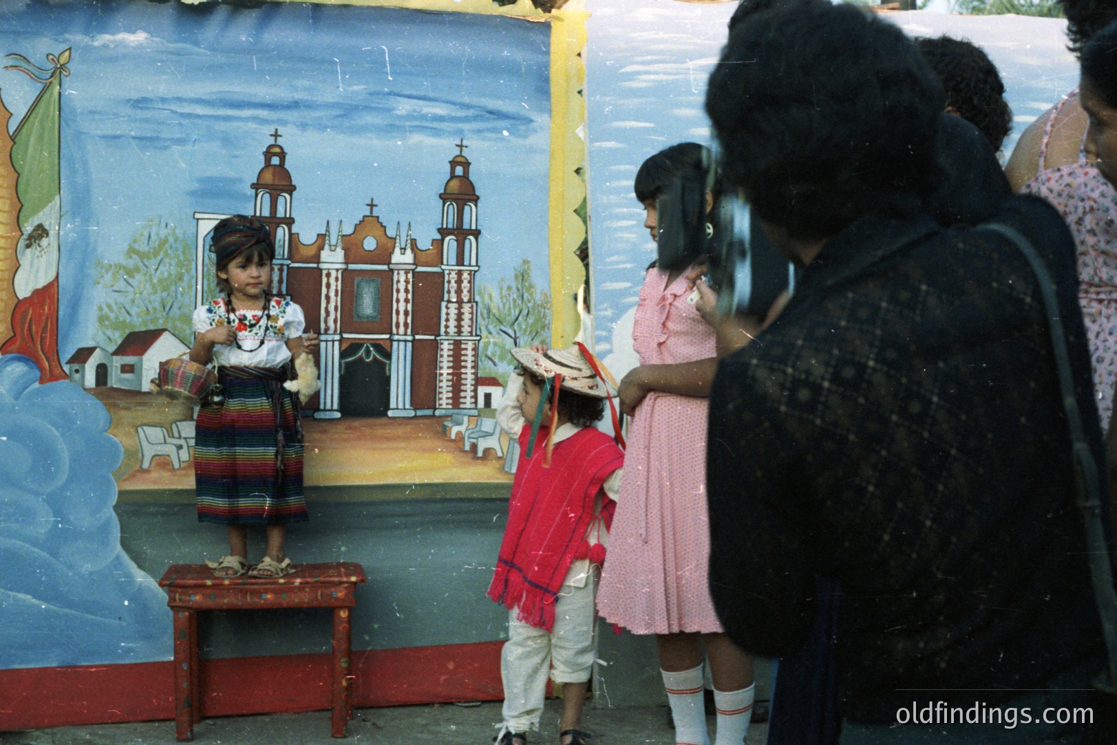 A young girl in traditional Mexican folk dress poses in front of a painted mural depicting a church, likely for a staged photograph. An adult holds a camera, capturing the scene with two children observing. The mural’s style suggests folk art. Likely 1970s or 80s.