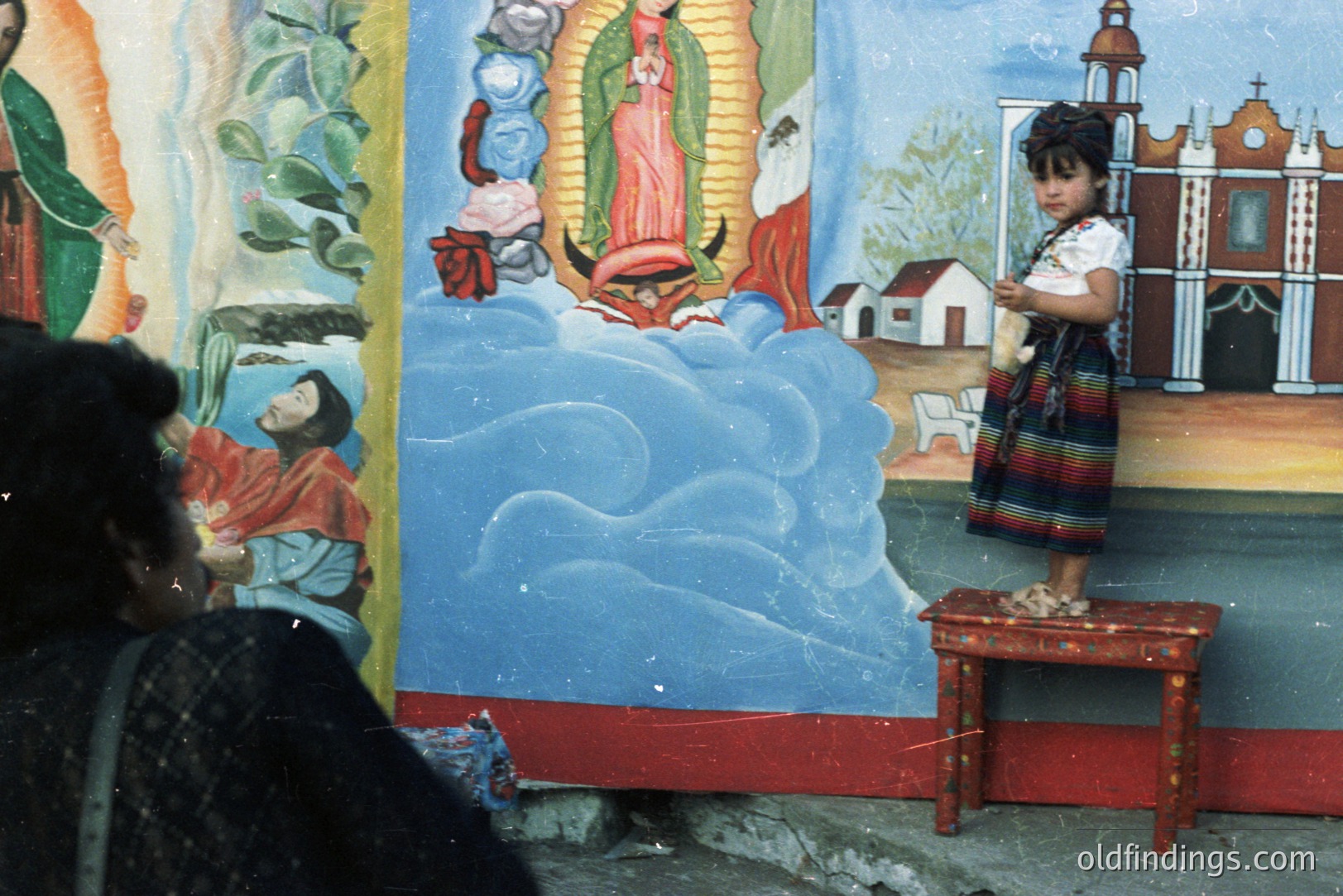 A young girl in traditional clothing stands on a small table before a vibrant mural depicting the Virgin Mary and a church facade. The mural’s style suggests a folk art or devotional image common in Mexican communities. Likely taken in the 1970s or 80s, the image captures a cultural moment.
