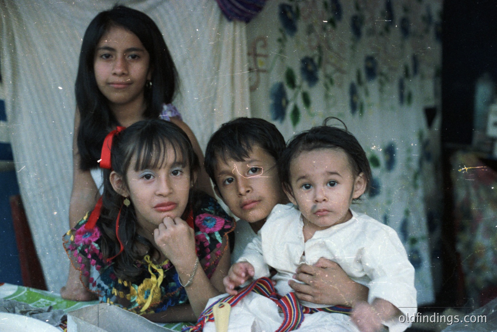 A poignant portrait of three children, seemingly indigenous, set against a patterned fabric backdrop. A young girl in a vibrant floral dress gestures thoughtfully, while two boys, one holding a toddler, gaze directly at the camera. Appears to be a candid, possibly documentary-style image.
