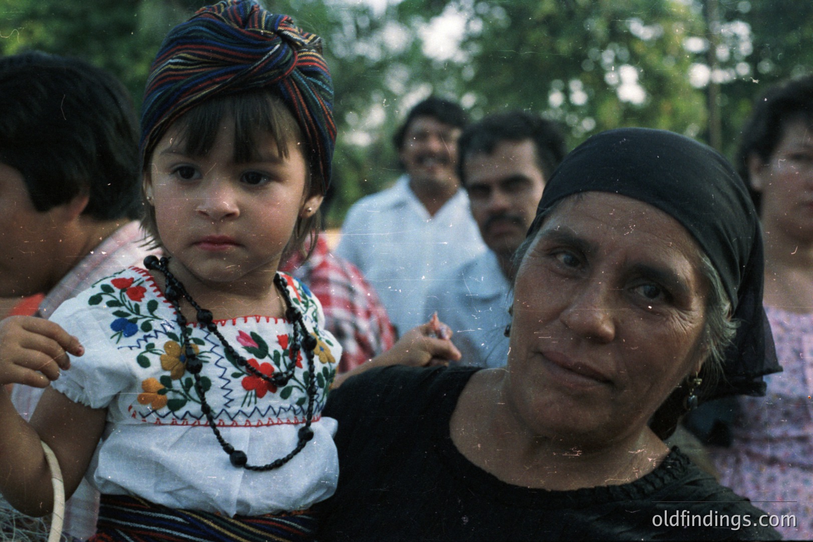A young girl in traditional embroidered clothing rests on a woman’s shoulders. The woman wears a dark headscarf. Background figures in casual dress suggest a community gathering. Likely Mexico, circa 1970s, based on clothing and photographic style. Cultural documentation; potential for design reference.
