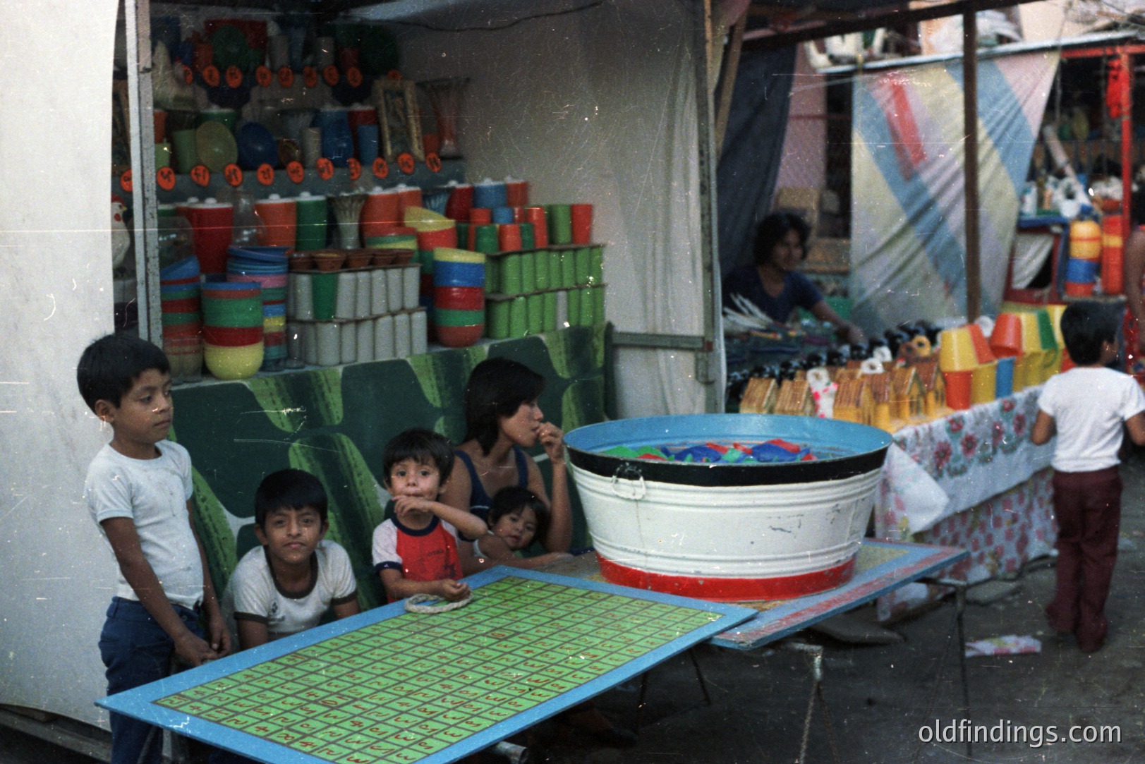 A vibrant street scene captures children near a vendor's stall overflowing with colorful plastic containers. The stall displays wares in green, red, and yellow. A large galvanized tub holds water; a vendor is partially visible. Likely a market or roadside shop. Likely 1970s.
