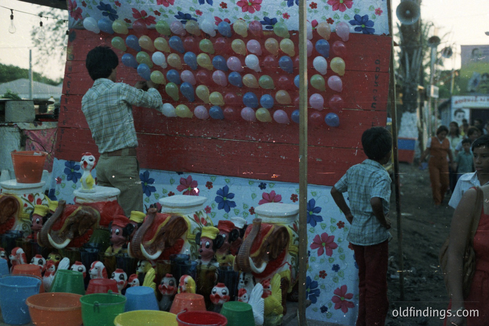 A vibrant, makeshift carnival game display featuring numerous balloons affixed to a floral-patterned backdrop. Two figures assist with balloon placement; a small crowd observes. Colorful plastic buckets and toys are arranged at the base. Likely a rural fair or community event.