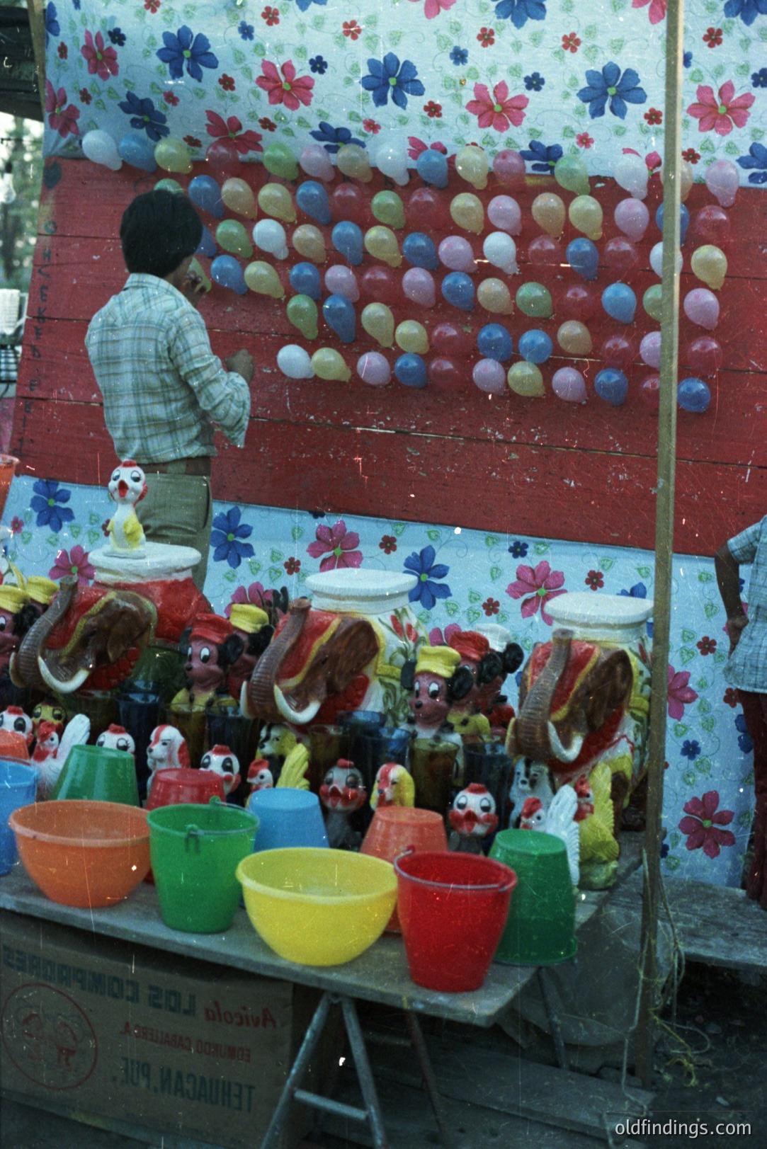 A vendor stands near a colorful display of ceramic rooster figurines and plastic buckets, set against a floral-patterned backdrop with numerous balloons affixed. The scene appears to be an informal marketplace or fair. Likely 1970s or 80s based on style. Demonstrates local craft & trade.