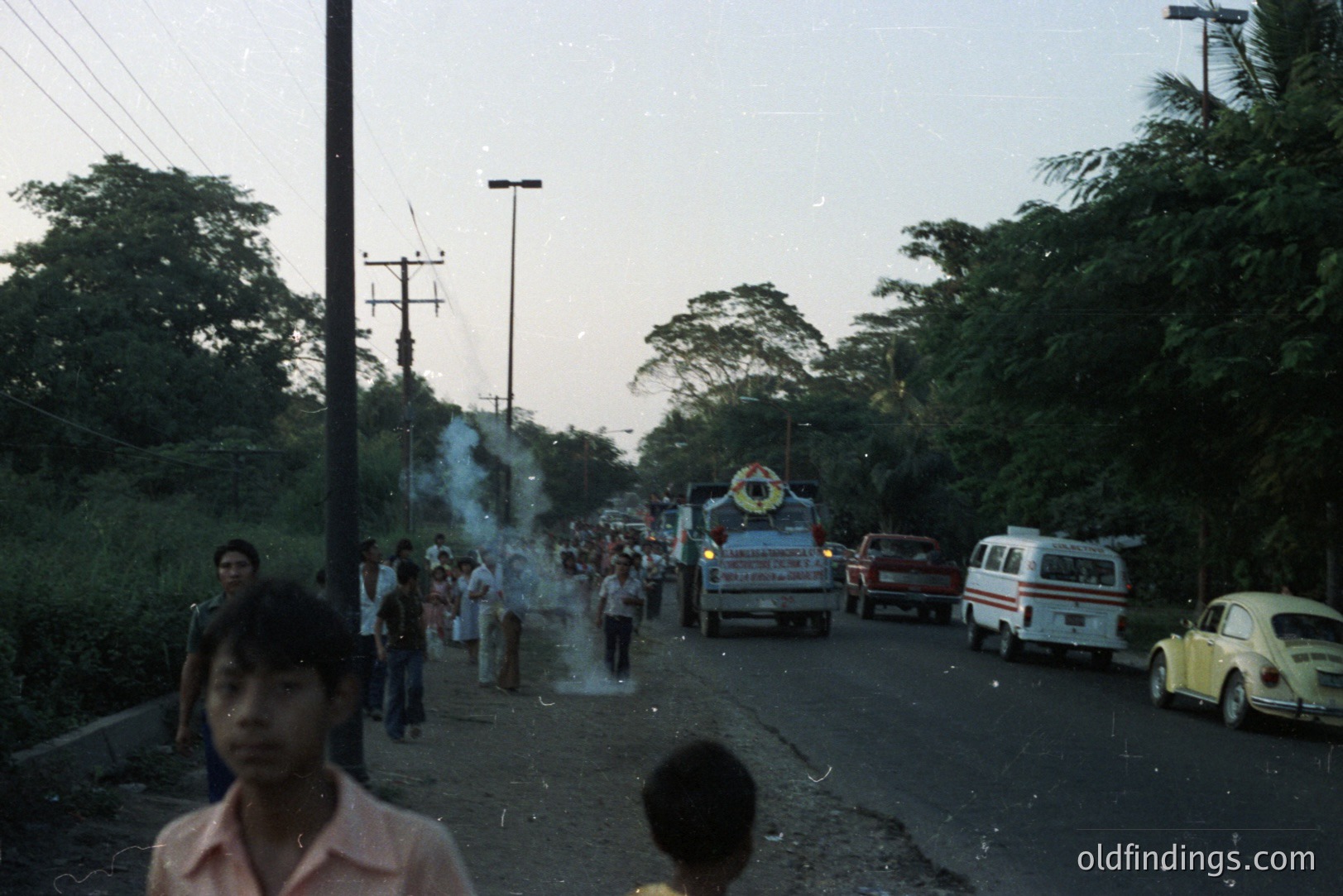 Street scene featuring a parade with a large decorated float, vehicles, and onlookers. Appears to be a rural town, possibly Southeast Asia, likely 1970s based on clothing and vehicles. A VW Beetle is prominently visible. The image shows a slice of local life and community celebration.