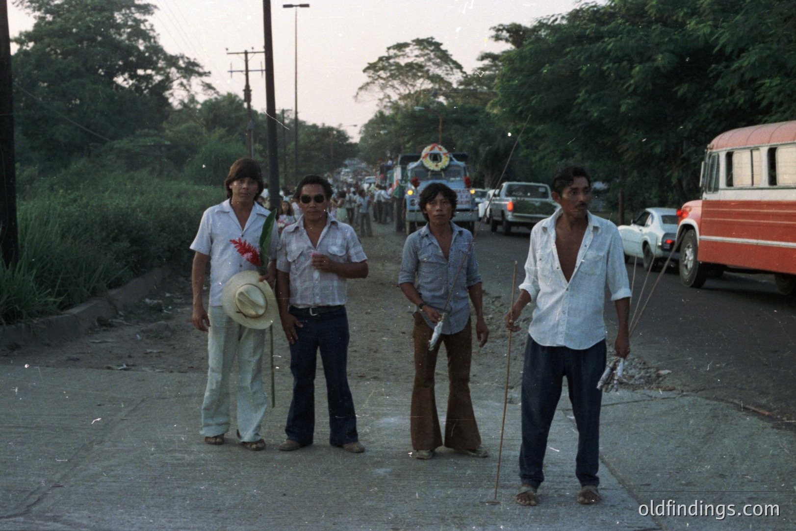 Four men stand on a paved road, appearing casually posed. They wear 1970s-era fashion—bell-bottoms, open shirts, and wide collars—suggesting a leisure or vacation setting. A vintage bus and various cars are visible in the background. Photograph likely depicts a coastal town.