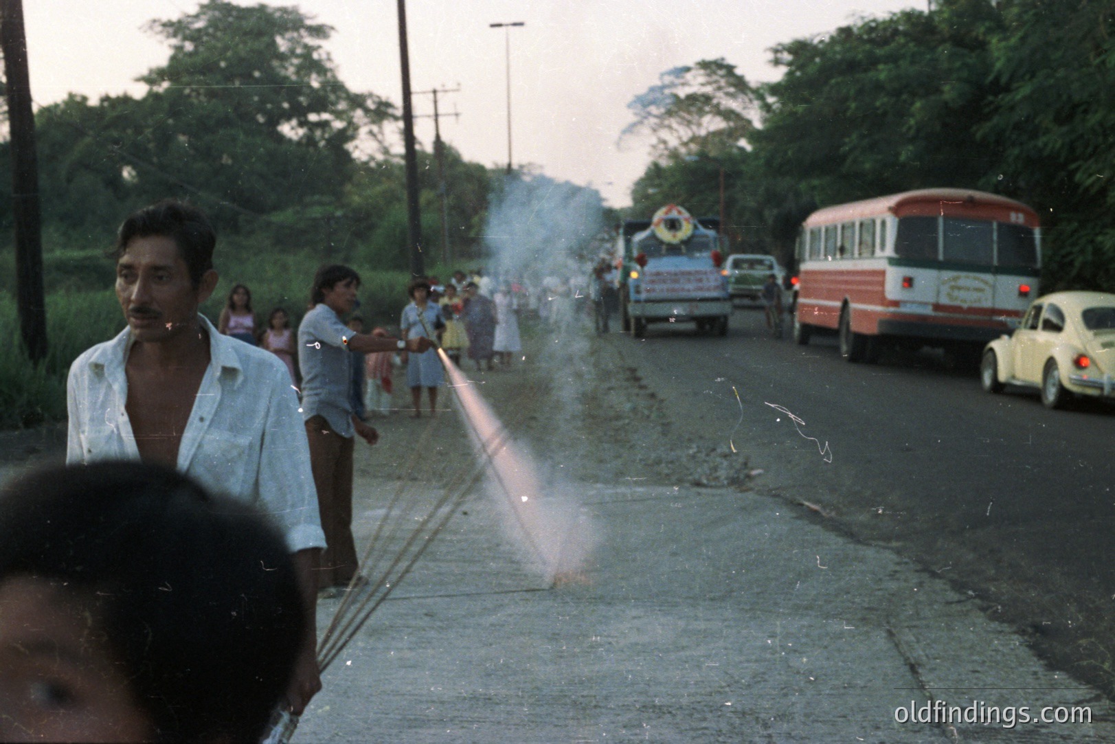 A roadside scene depicts men lighting fireworks on an asphalt road. Children and onlookers watch as the smoke rises. A bus, trucks, and a vintage Volkswagen Beetle are visible in the background, indicative of a rural location. Appears to be a casual, candid moment.