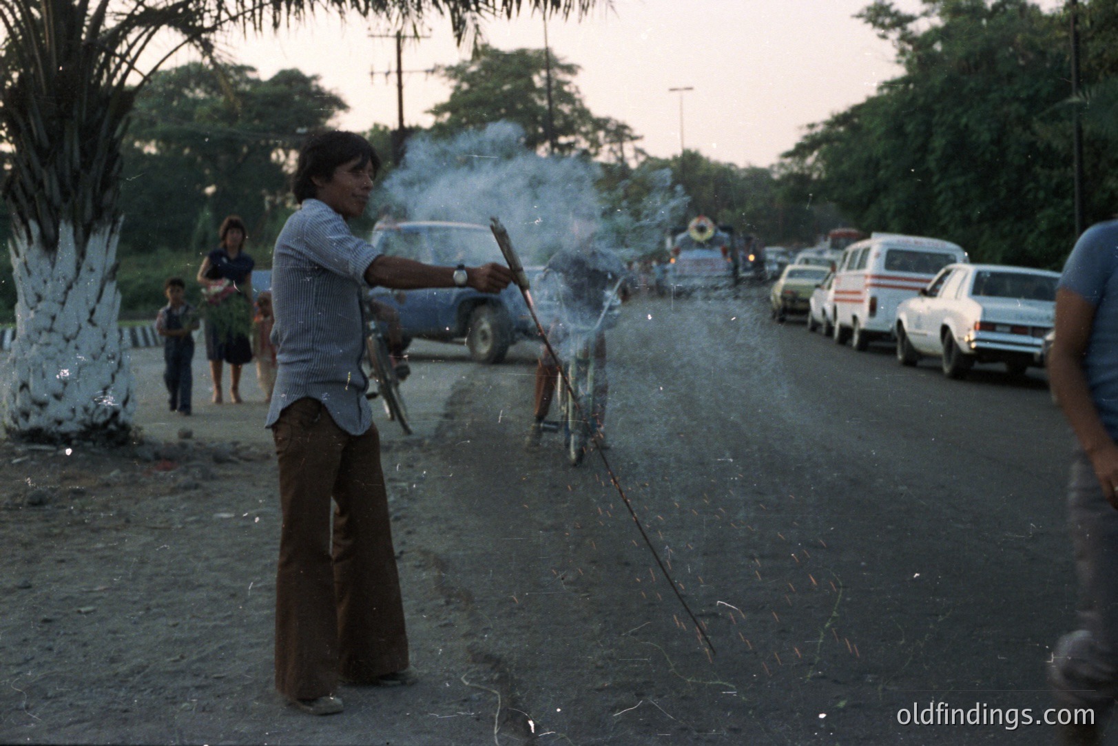 A man in flared trousers and a button-down shirt ignites a string of firecrackers on a dusty road. A van, several cars, and onlookers are present. The scene evokes a festive, possibly rural, 1970s setting. Likely documenting a cultural tradition or celebration.