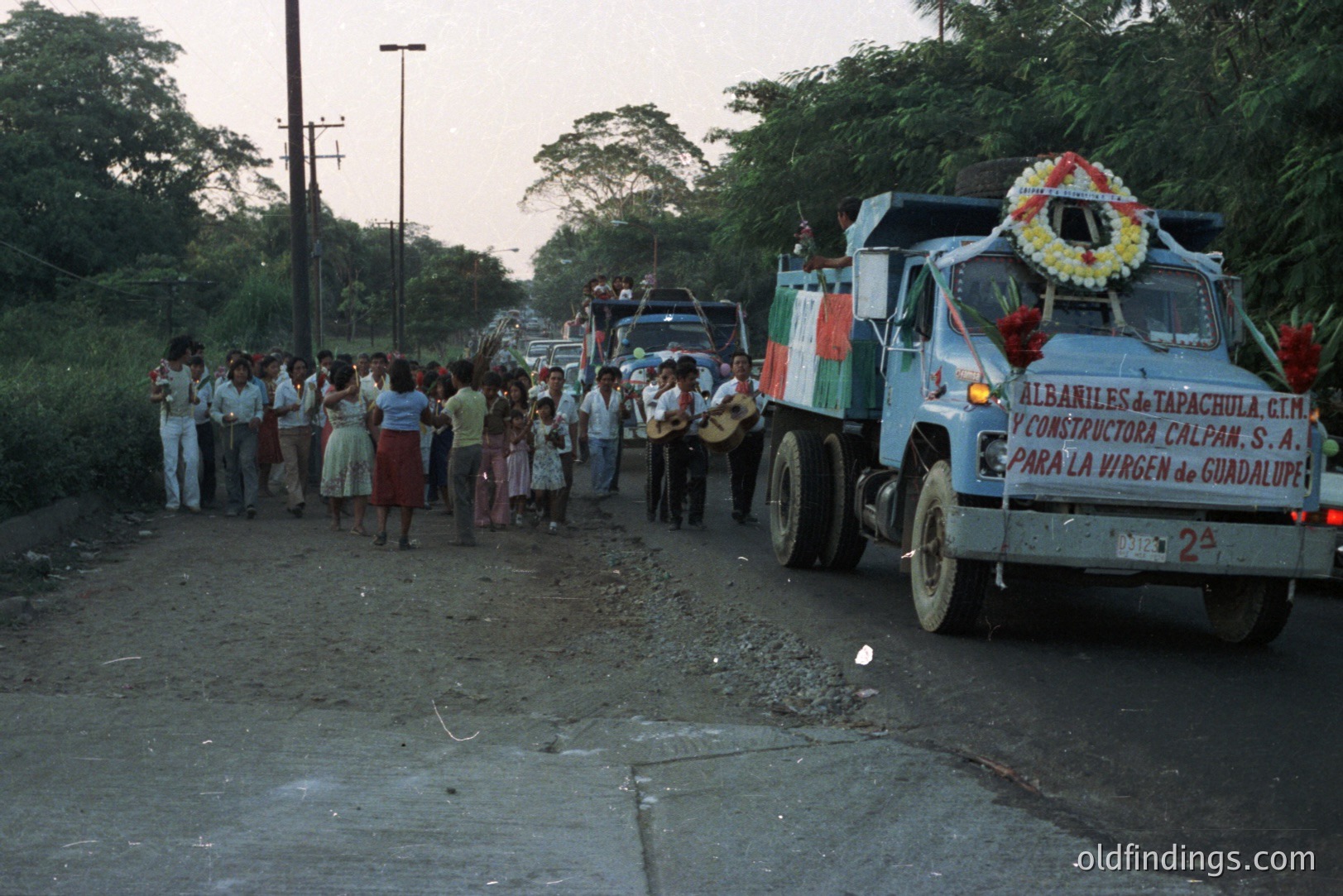 A celebratory procession unfolds on a rural road. A light blue dump truck, adorned with flowers, leads a group of people, some playing musical instruments, while others watch or participate. Likely a local festival or religious observance. Appears to be Mexico, possibly 1970s.