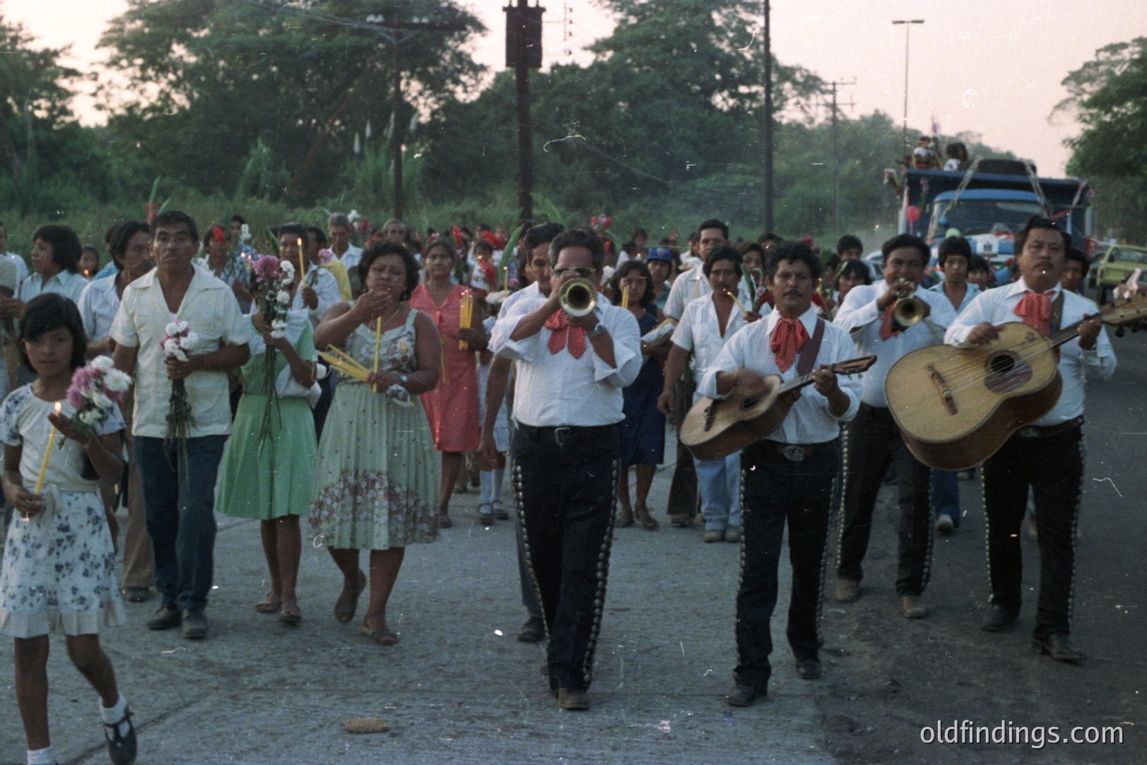 A vibrant street procession features a mariachi band in traditional white shirts and black embroidered pants. Attendees, many in colorful dresses, watch the parade. Likely a rural Mexican town, capturing festive local culture. The photograph's style suggests the 1970s or 80s.