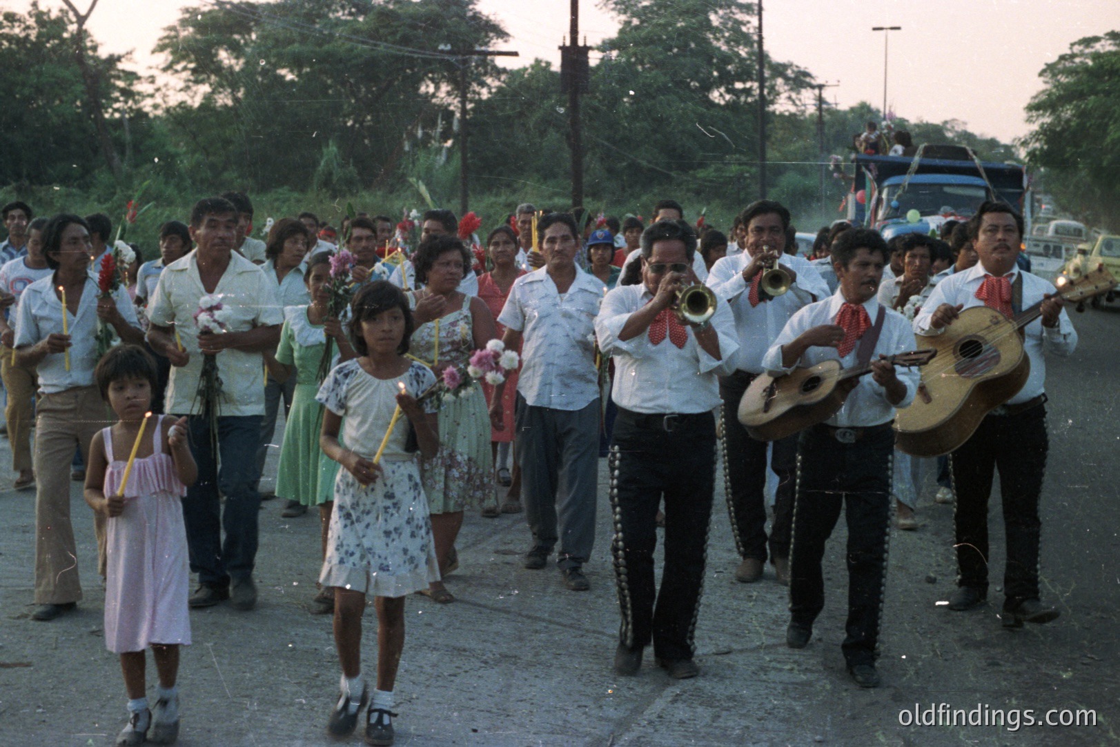 Procession featuring a musical ensemble and community members in traditional attire. Candle bearers lead the group, followed by musicians playing trumpets, guitars, and accordions. Likely a religious festival or local celebration. Appears to be rural Mexico, 1970s, based on style & photographic technique.