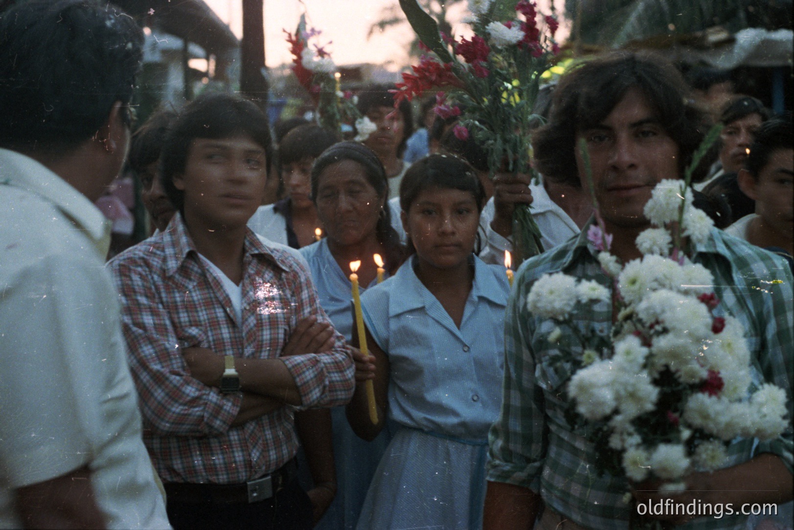 A community gathers during a religious procession. Individuals hold candles and bouquets of flowers. Clothing styles and hairstyles suggest a timeframe of the 1970s or 1980s, possibly Latin America. The photograph’s color palette hints at a tropical location. Image displays cultural traditions and community bonds.